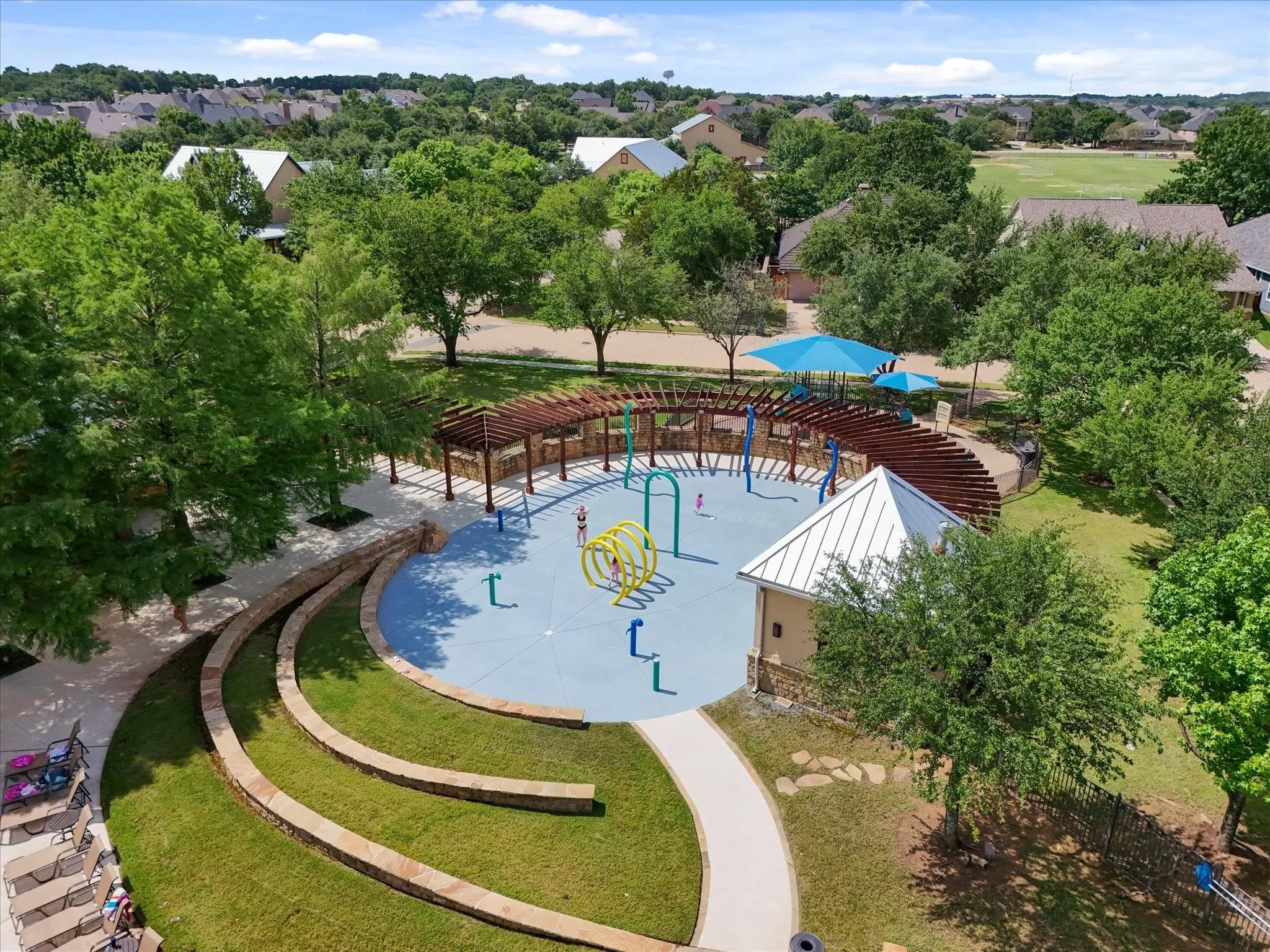 Community splash pad featuring interactive water elements, a shaded pergola, terraced seating, a utility building with a metal roof, and surrounding mature trees