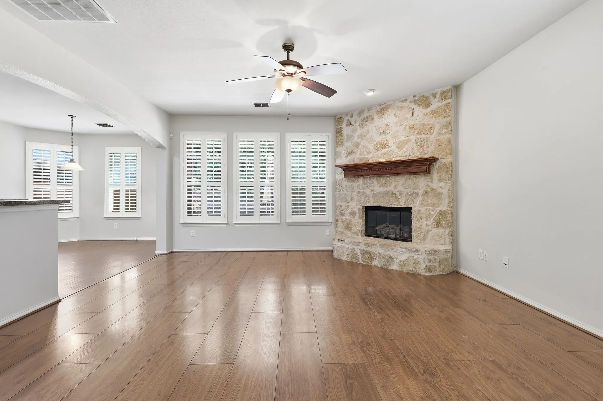 Wood-finish flooring, stone-clad fireplace with wood mantle, multiple windows with plantation shutters, ceiling fan, and pendant lighting