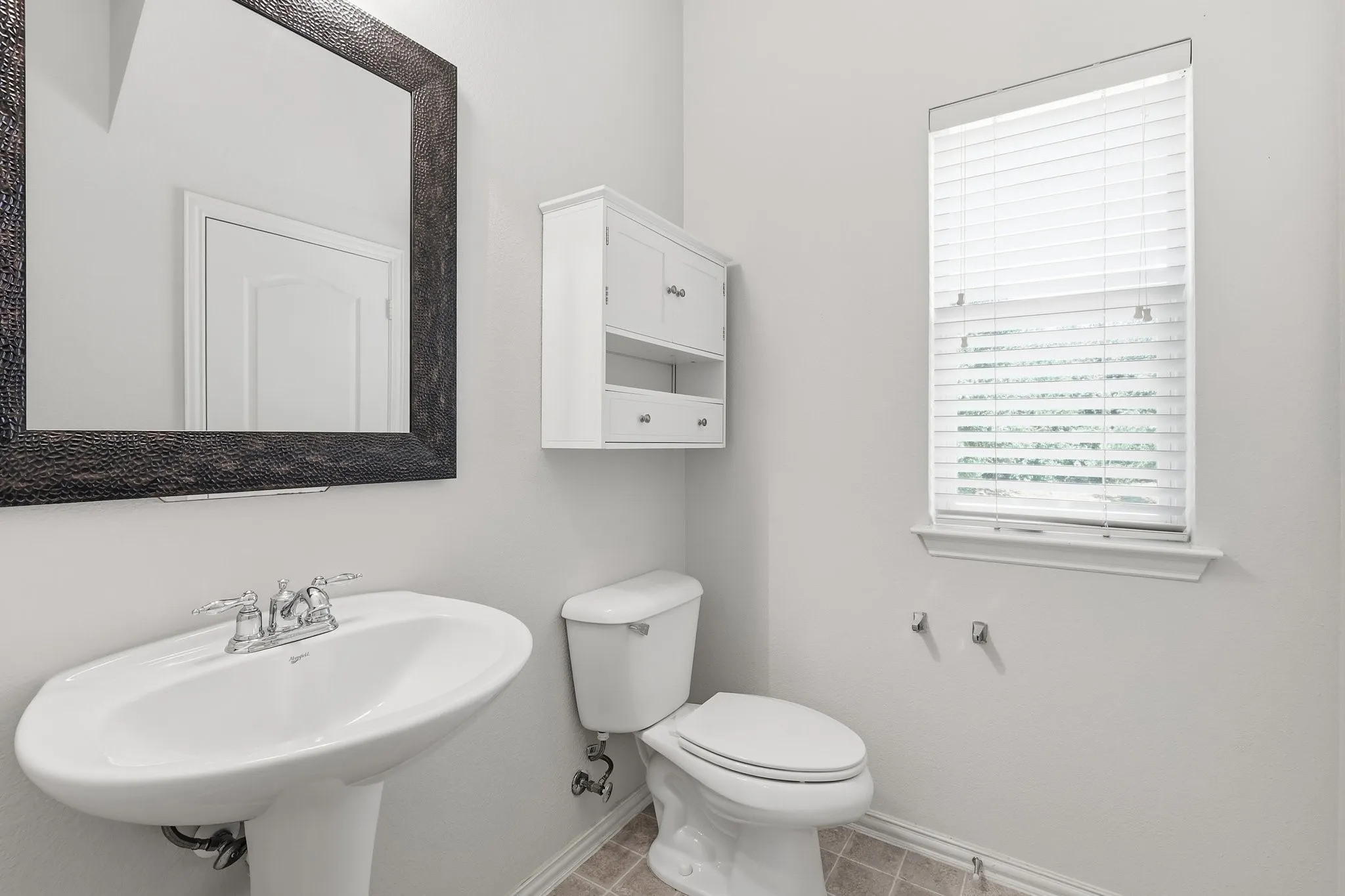 Pedestal sink with chrome fixtures, framed wall mirror, white wall-mounted cabinet, window with privacy blinds, and tile flooring