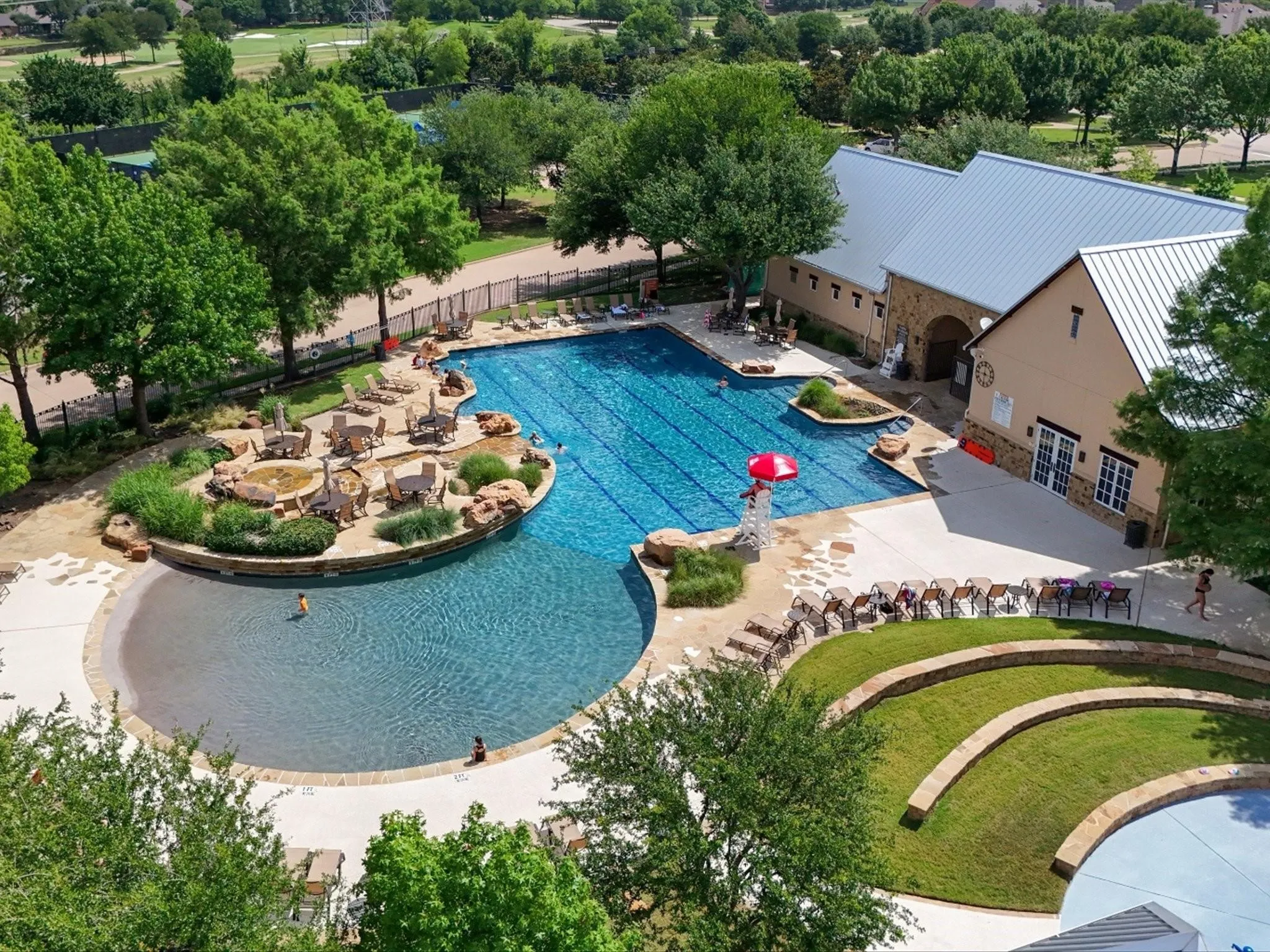 Expansive community pool area featuring a large swimming pool, a shallow entry pool, structured landscaping, and a clubhouse with a metal roof