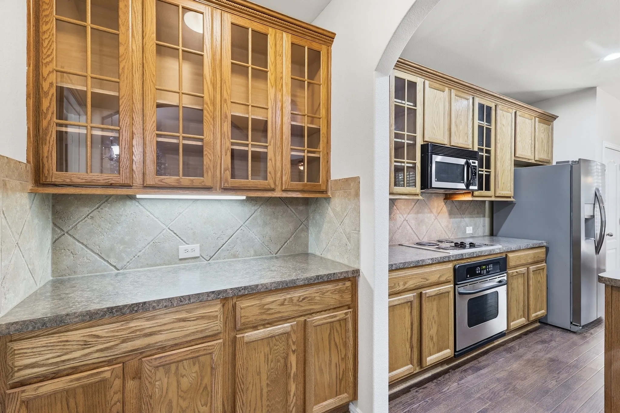 Wood-finish cabinetry with glass-front uppers, tiled backsplash, stainless steel appliances, and wood-finish flooring