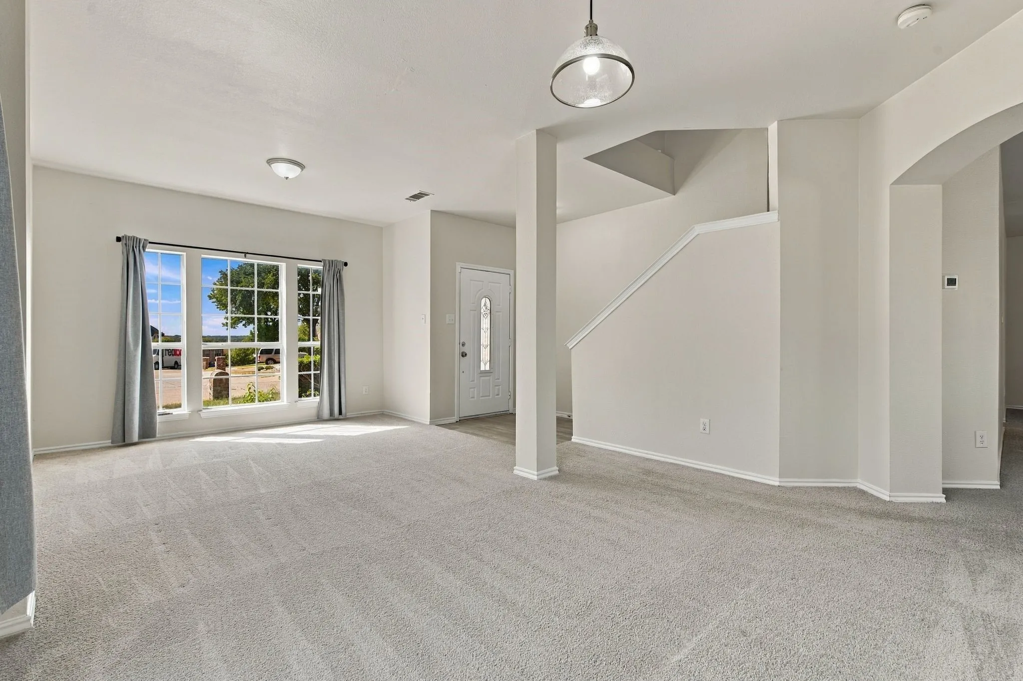Spacious room with light gray carpeting, a multi-pane window, and a white front door with decorative glass