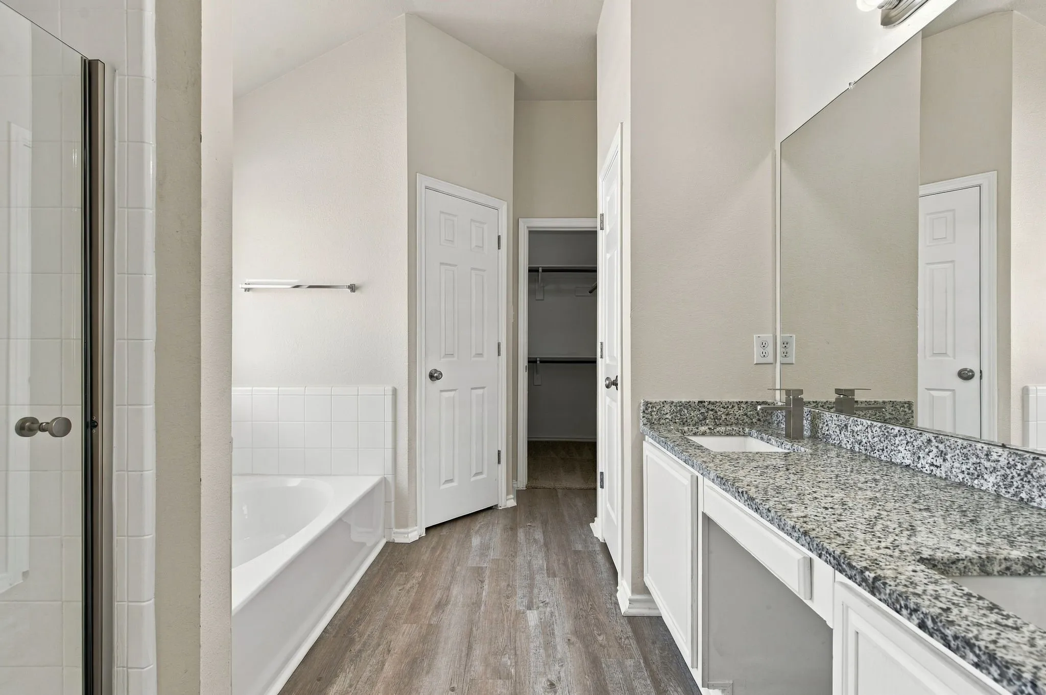 Dual vanity with stone countertop, white cabinetry, wood-finish flooring, and a tiled shower with glass enclosure