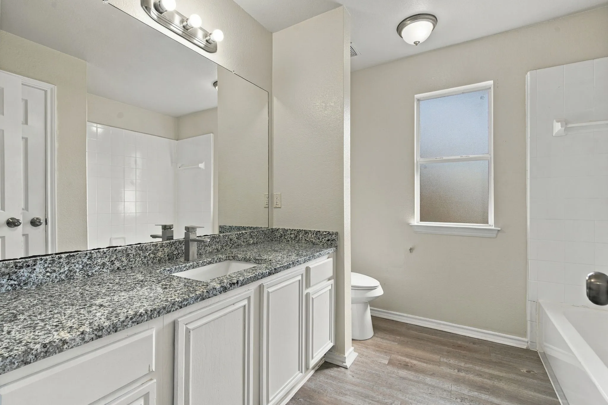 Vanity with white cabinetry and granite-style countertop, large vanity mirror, modern light fixture, wood-finish flooring, and a window