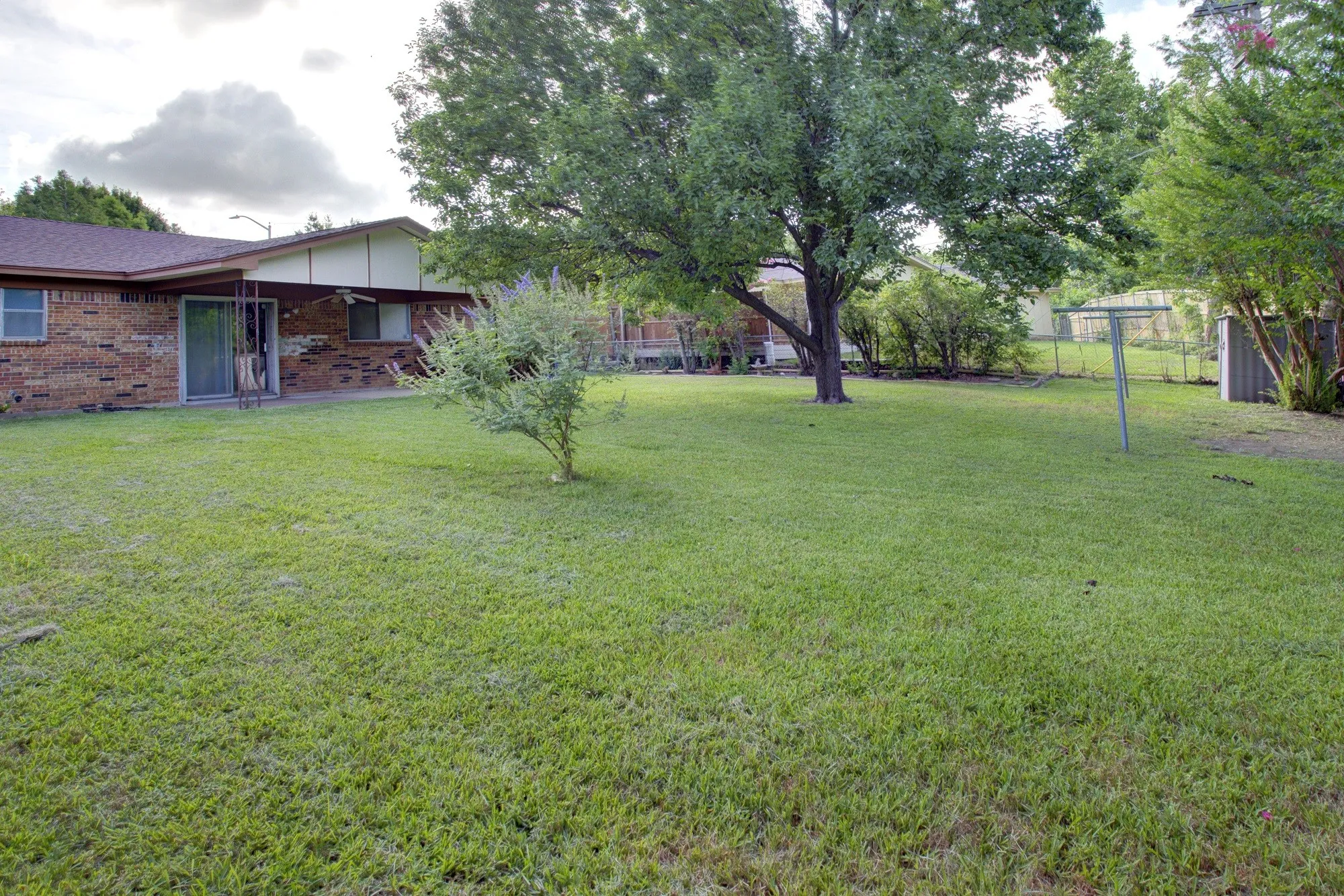 Expansive rear yard with mature trees, a brick-clad exterior, sliding glass door, and a utility shed
