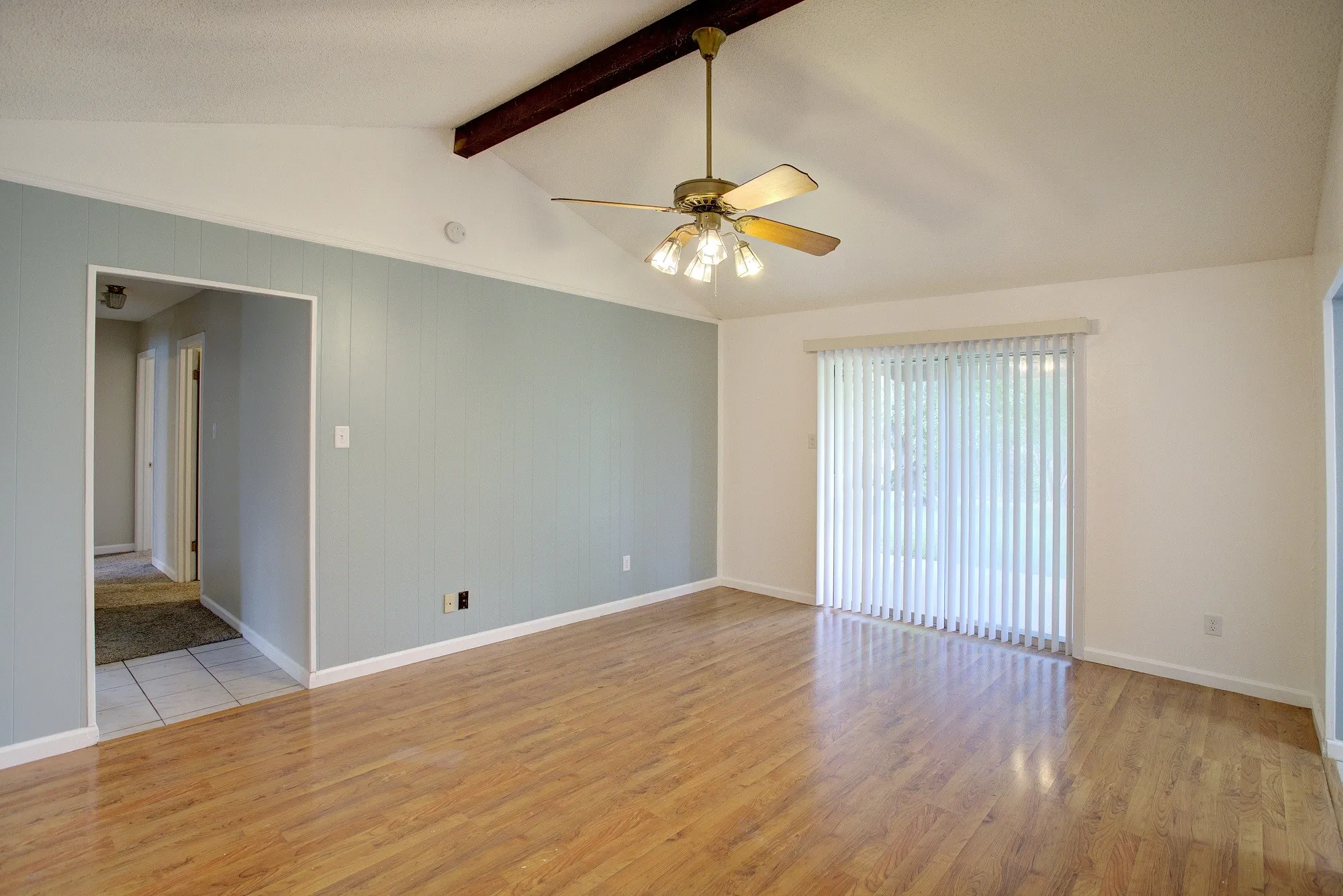 Vaulted ceiling with exposed wood beam, wood-finish flooring, ceiling fan with light kit, sliding glass door with vertical blinds, and vertical plank wall paneling