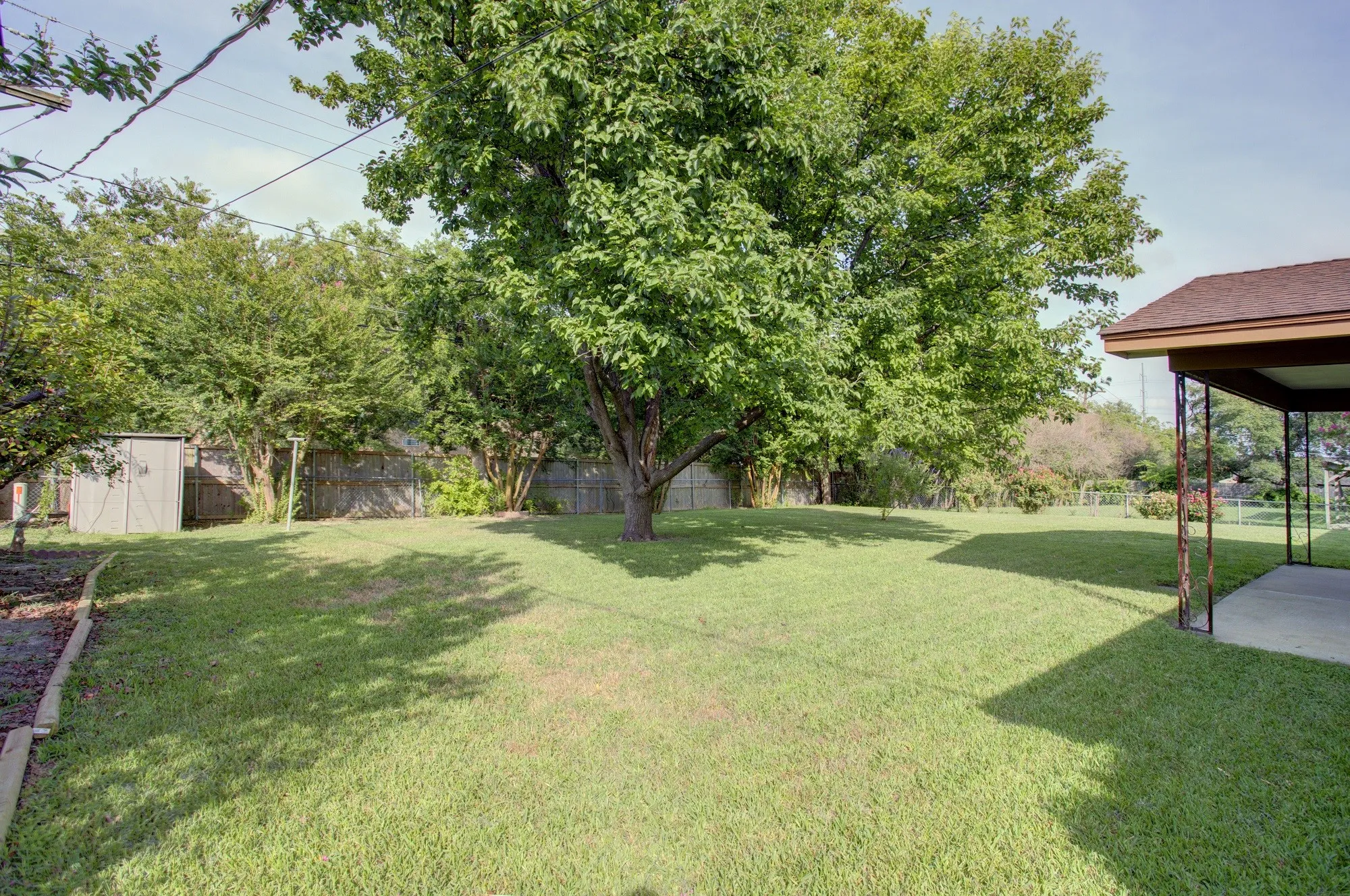 Expansive grassy yard with mature trees, a covered patio, and a utility shed
