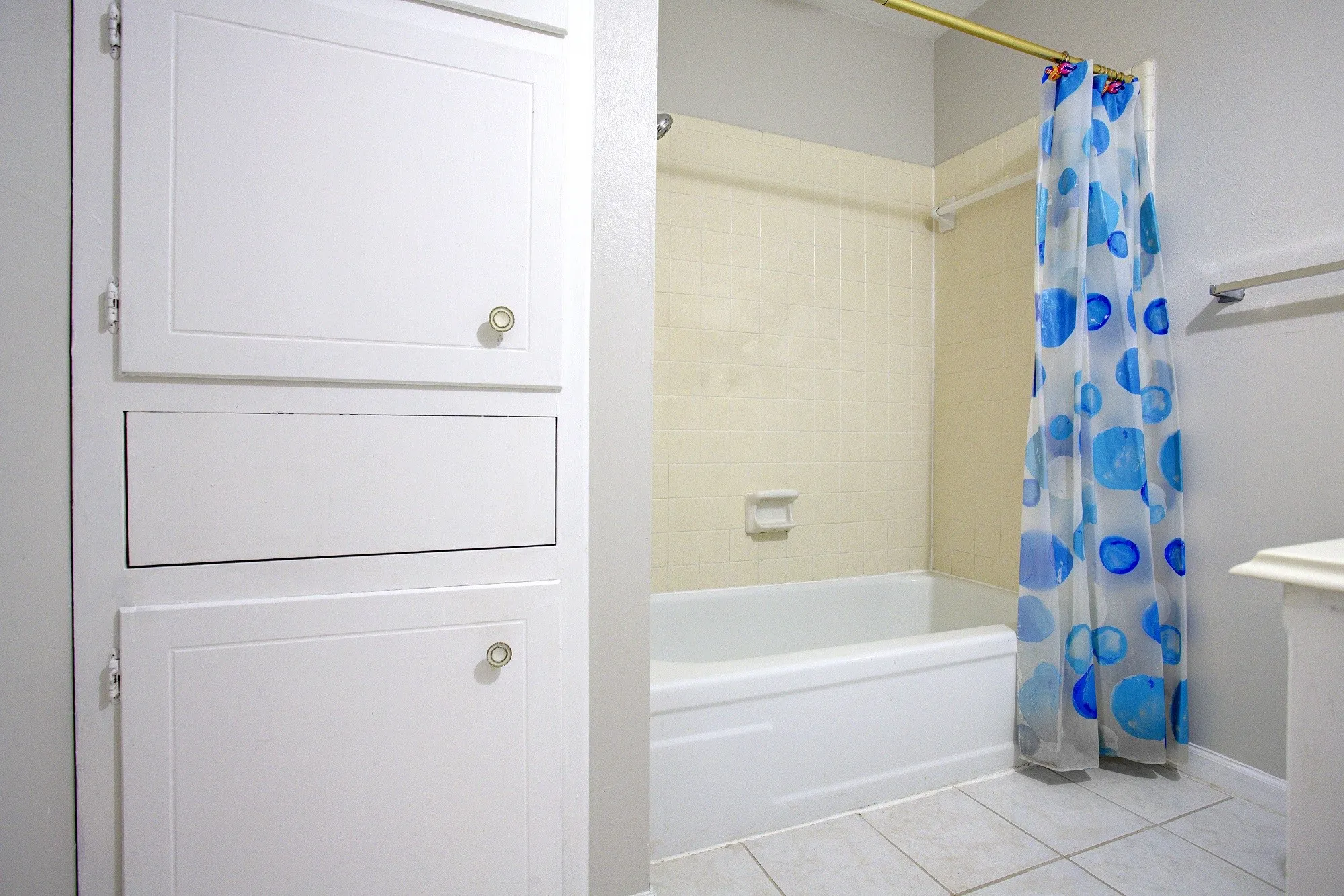 Built-in white cabinetry with gold-finish hardware, white bathtub with a tiled surround, and light-toned tile flooring