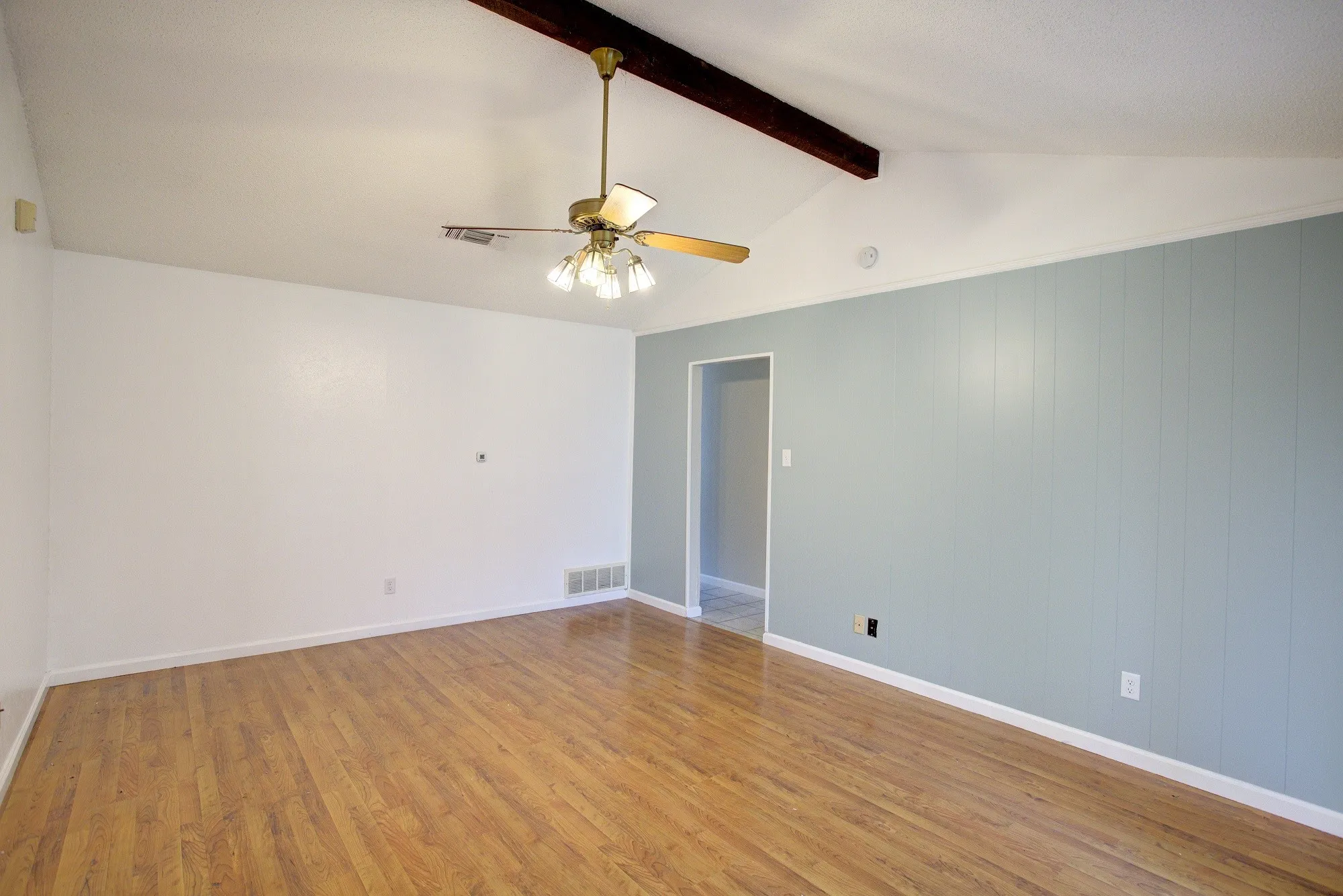 Wood-finish flooring, vaulted ceiling, exposed beam, ceiling fan with light fixture, and paneled accent wall