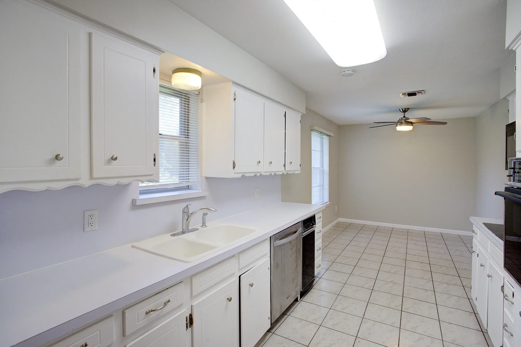 White cabinetry, dual basin sink with chrome faucet, stainless steel dishwasher, tile flooring, and a ceiling fan with light fixture