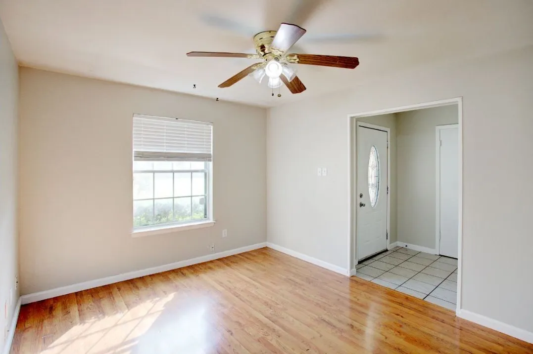 Warm wood-finish flooring, a window with horizontal blinds, and a ceiling fan with integrated lighting