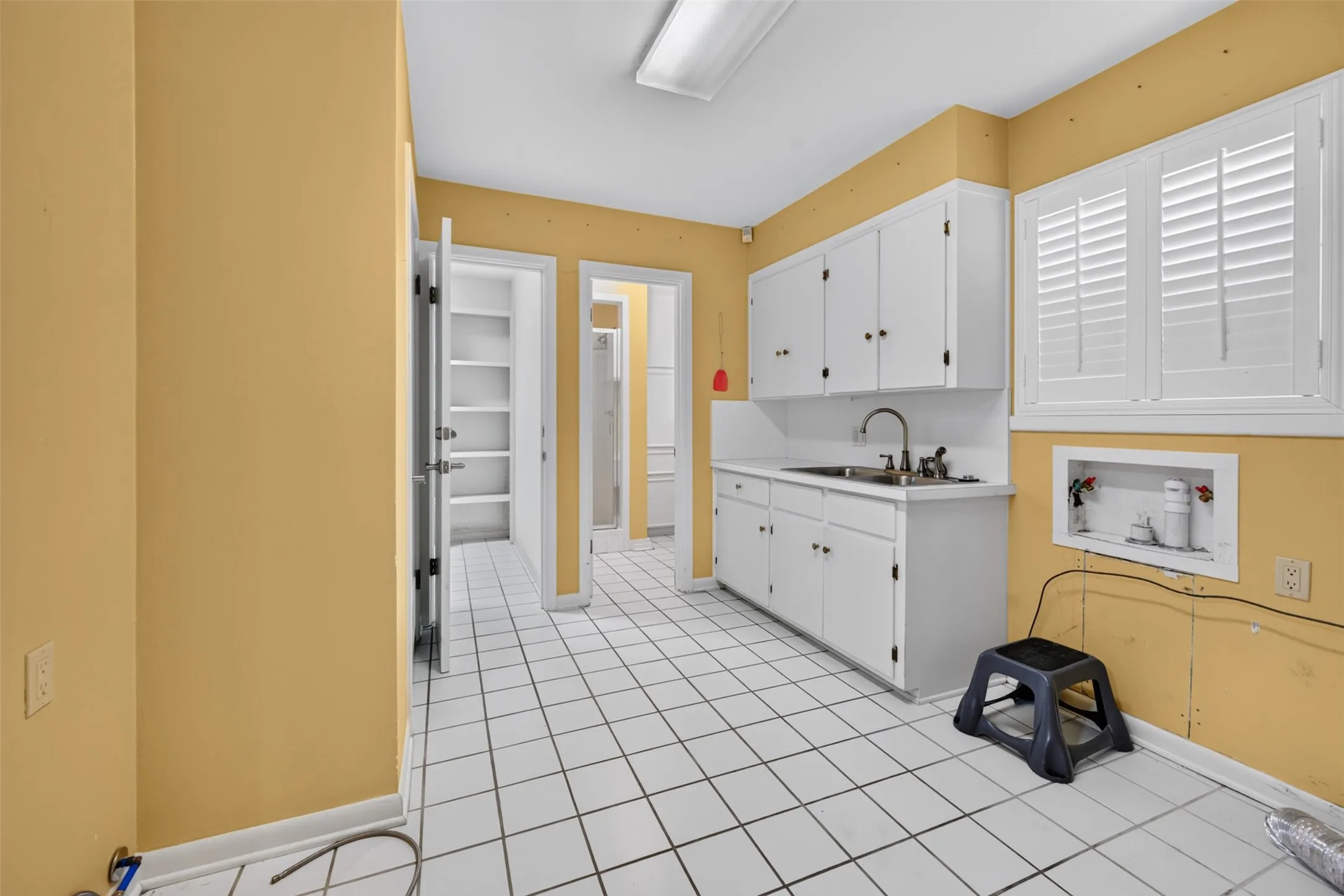 White square tile flooring, white cabinetry, stainless steel sink, window with white plantation shutters, built-in shelving