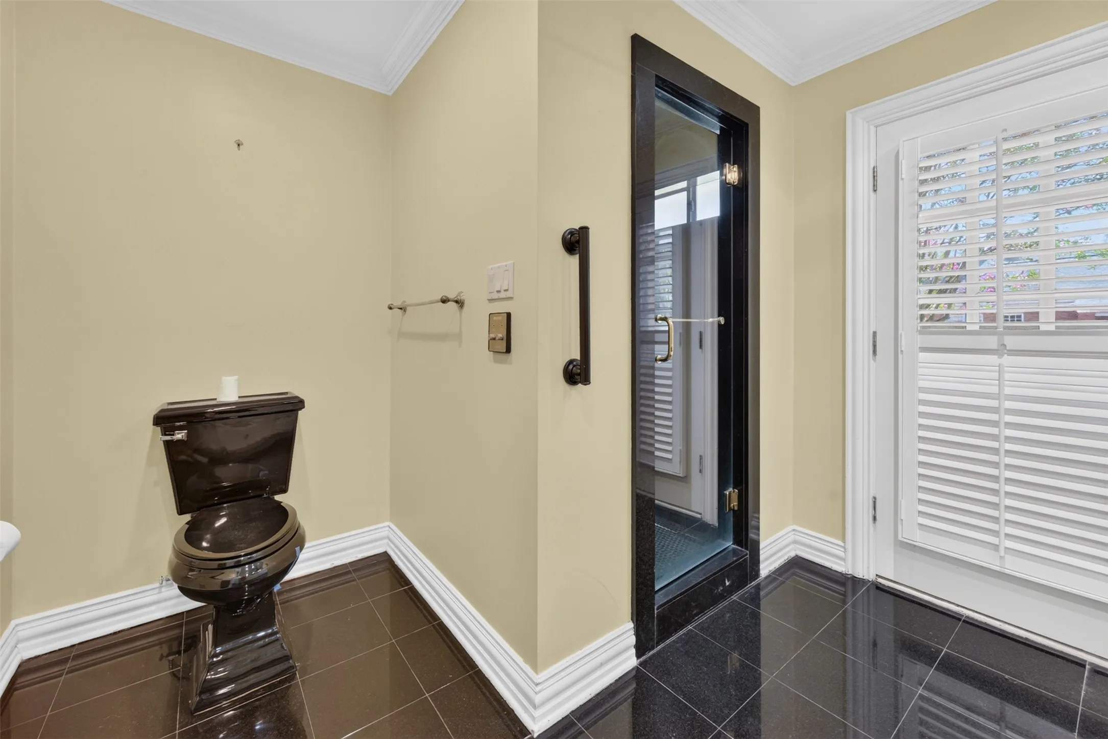 Black tile flooring, white crown molding, black toilet, glass-paneled door, and a white plantation shutter window