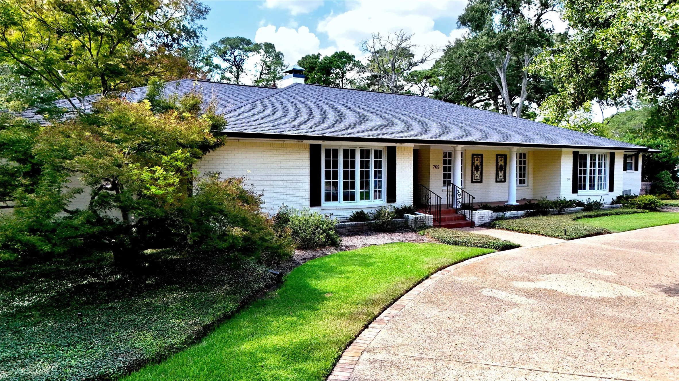 Brick exterior, dark roof, front porch with columns, mature landscaping, and a paved driveway