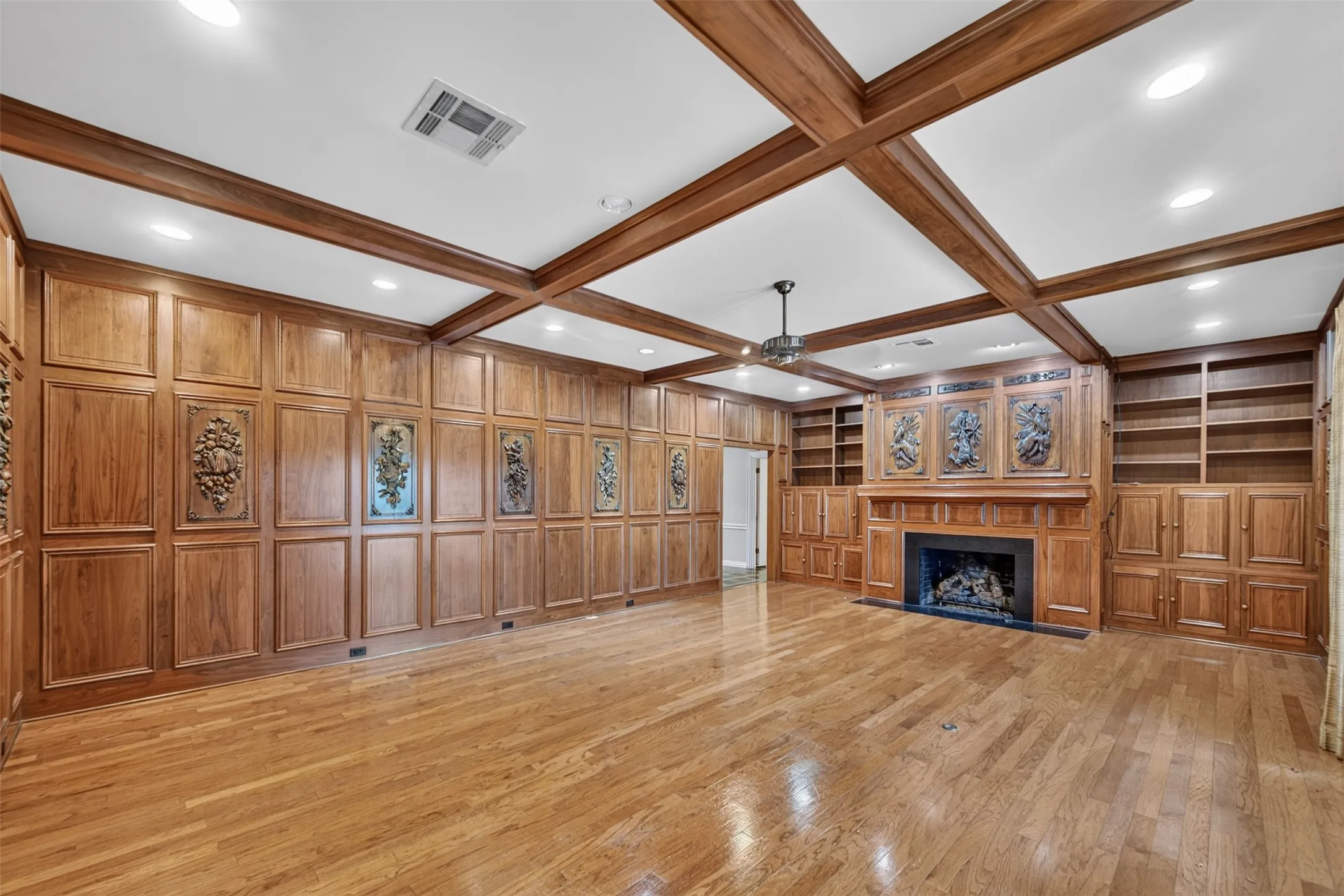 Wood-paneled room featuring a coffered ceiling, a fireplace with a stone surround, built-in shelving, and wood flooring