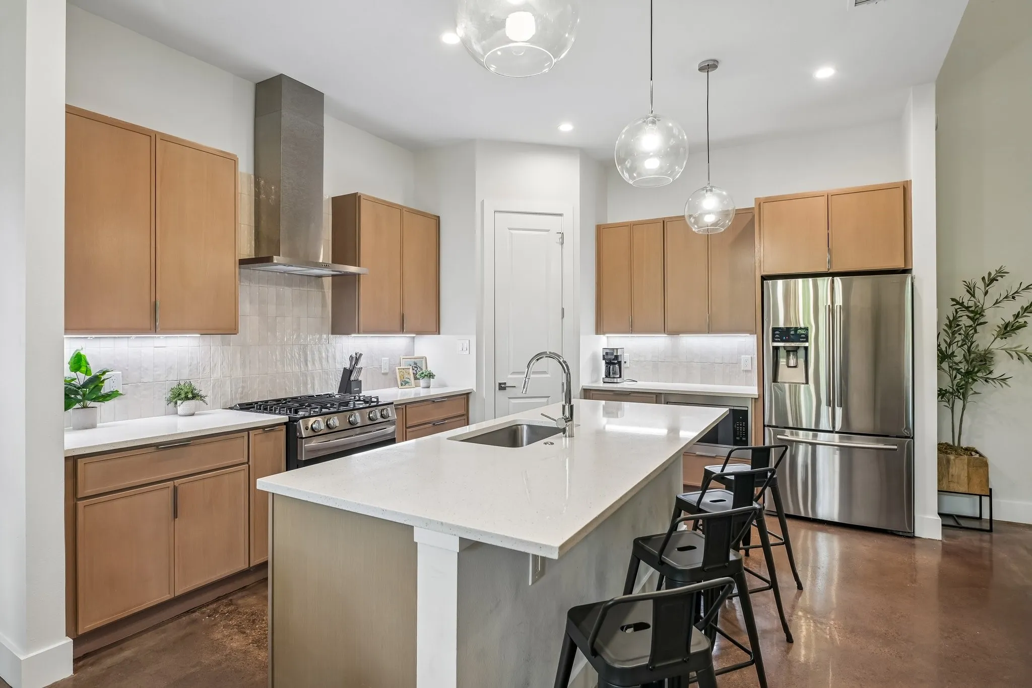 Kitchen featuring stainless steel appliances, a center island with sink, light wood-finish cabinetry, a gas range with a stainless steel vent hood, and pendant lighting