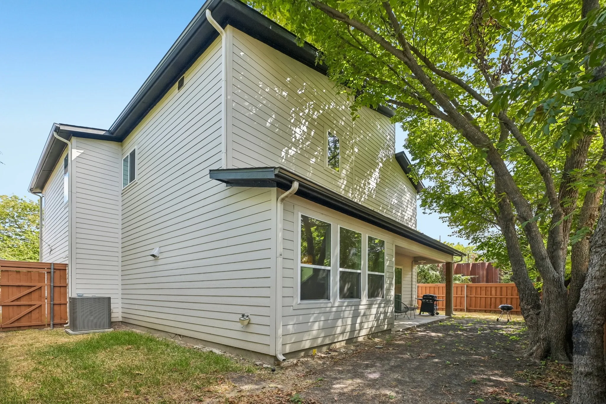 Rear exterior featuring light-colored siding, a covered patio, multiple windows, and mature trees providing shade