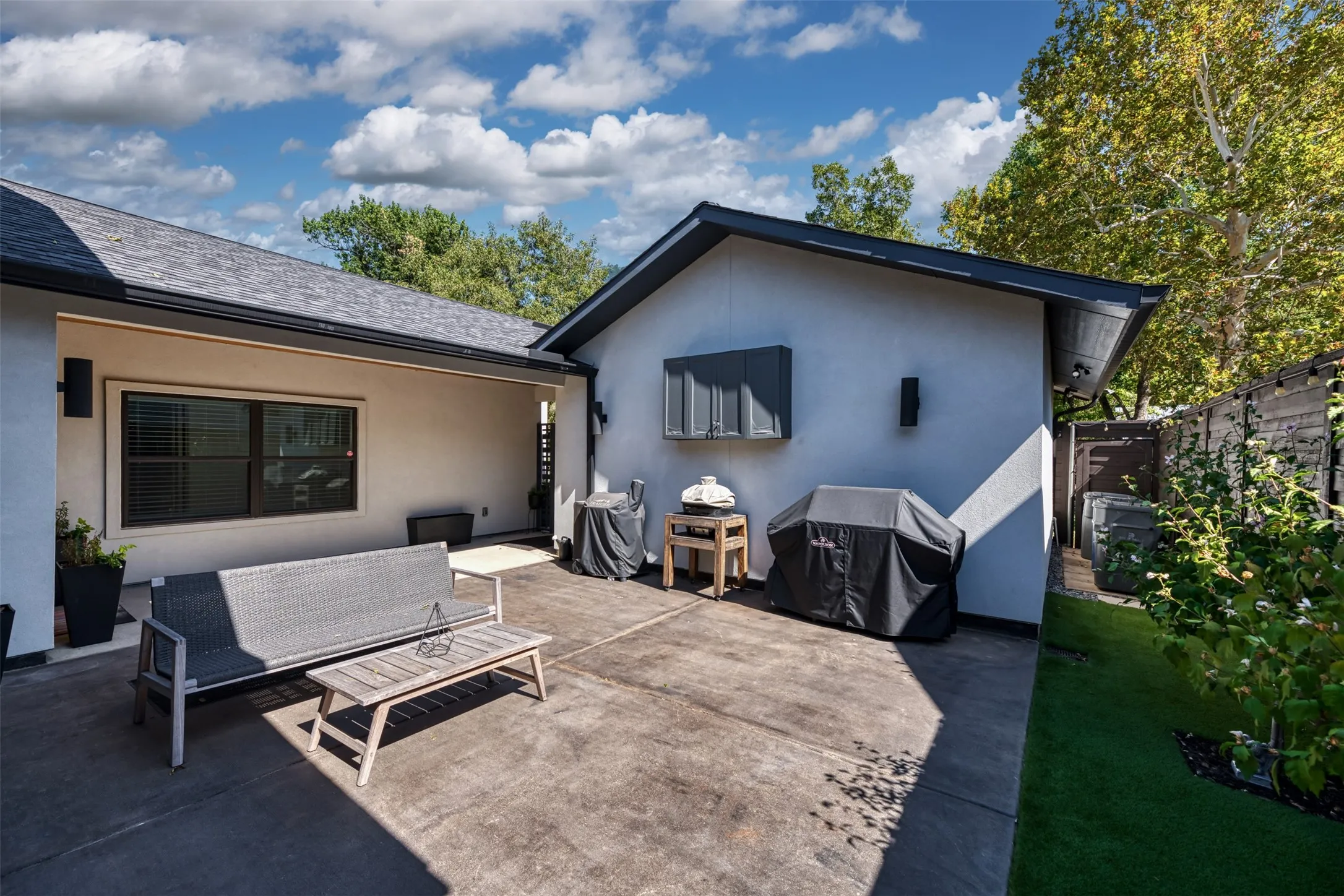 Paved patio area, stucco exterior, dark window trim, dark roof shingles, and established landscaping