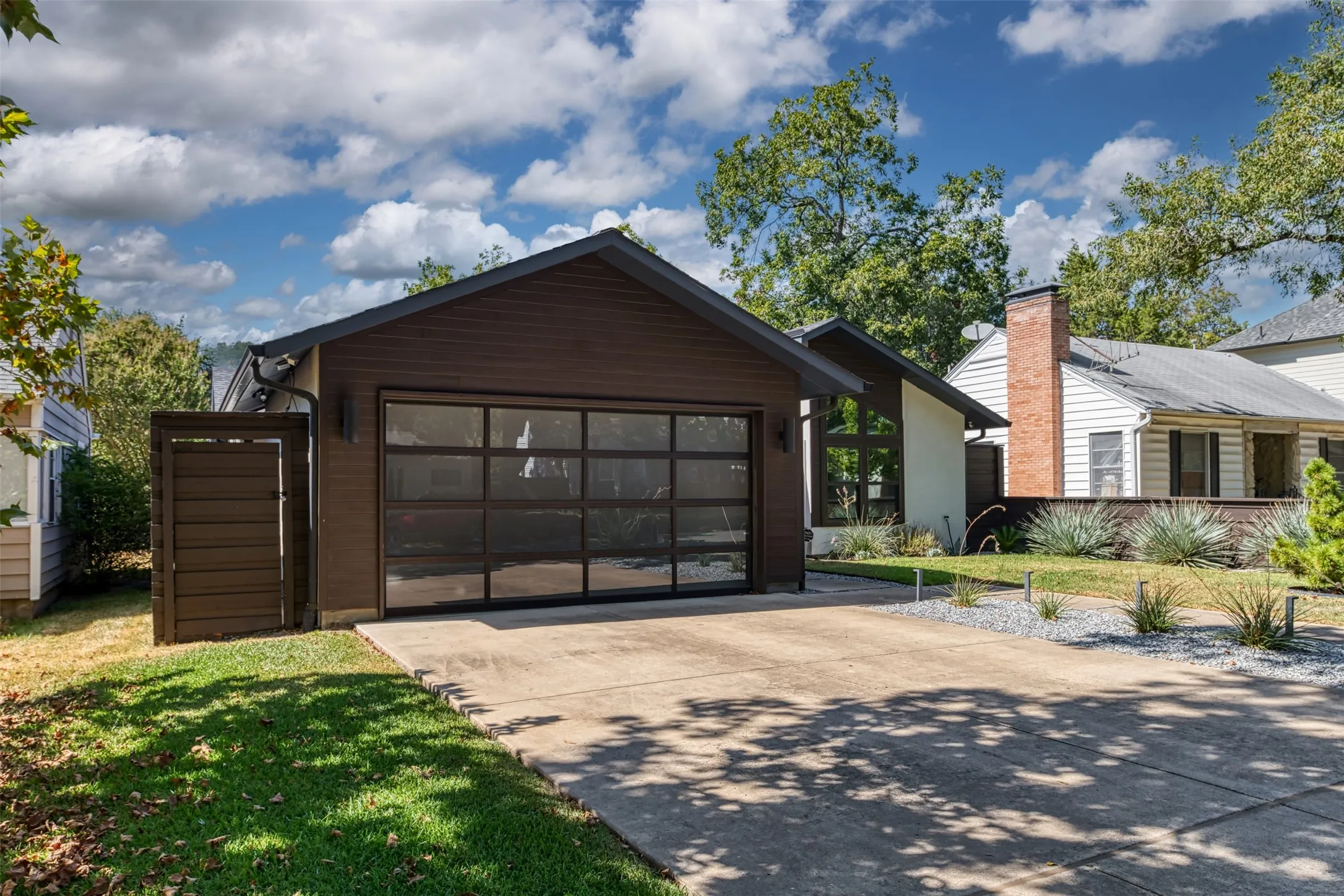 Concrete driveway, modern glass-paneled garage door, dark wood-finish siding, sloped roofline, and landscaped front yard with decorative rocks