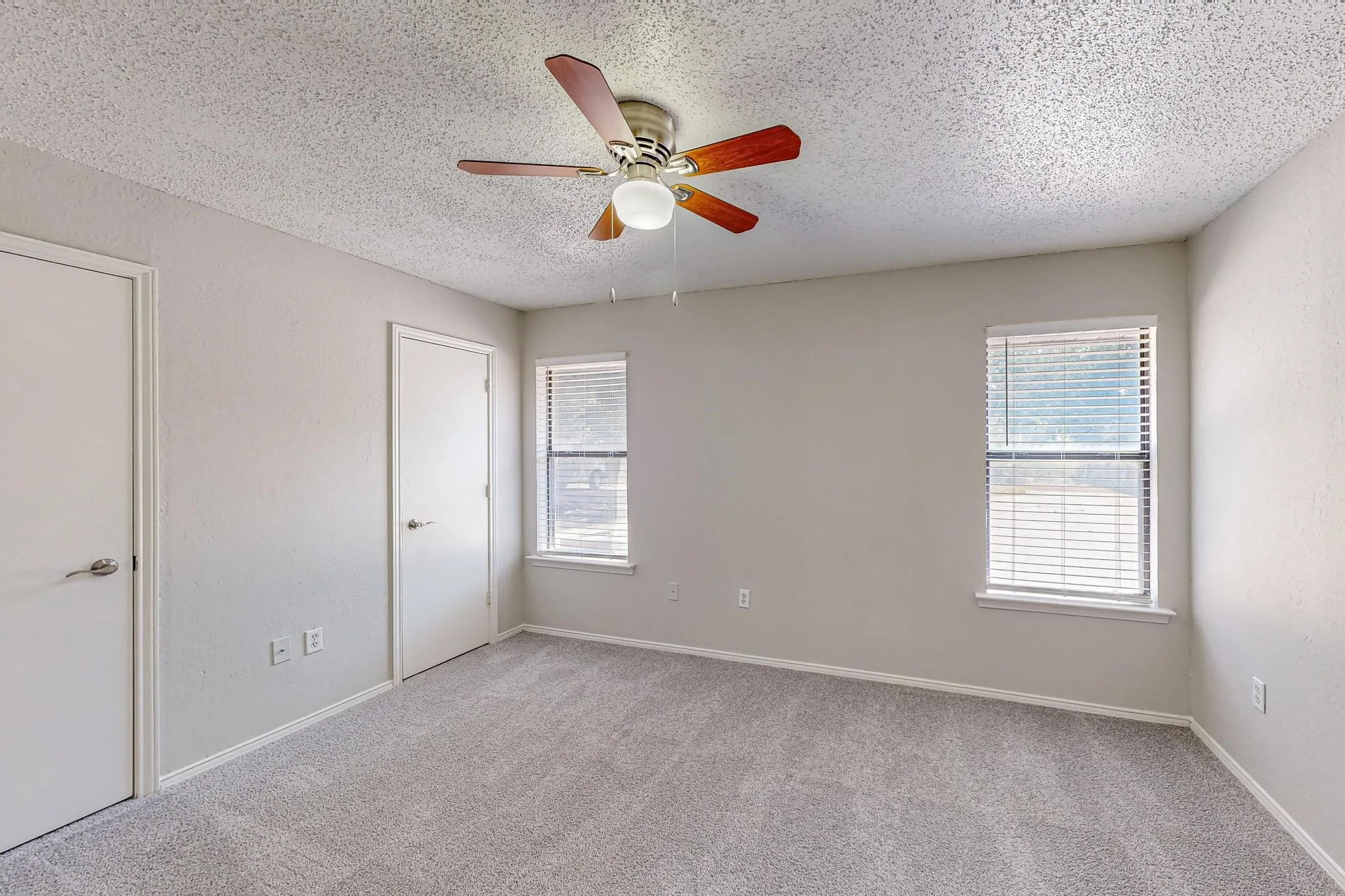 Spacious room featuring neutral carpeting, two windows with blinds, two interior doors, and a ceiling fan with integrated lighting