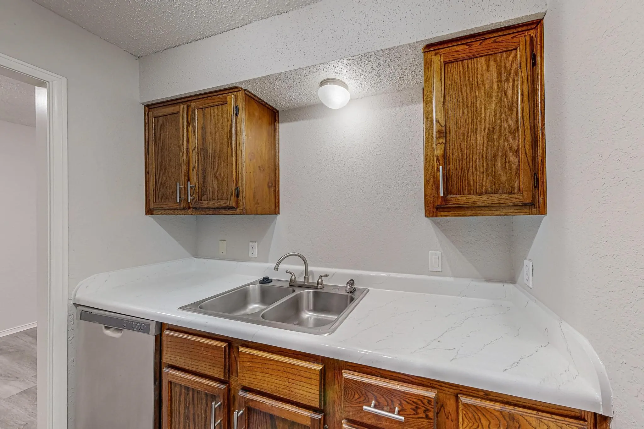White patterned countertop, double basin stainless steel sink with chrome faucet, stainless steel dishwasher, and wood-finish cabinetry