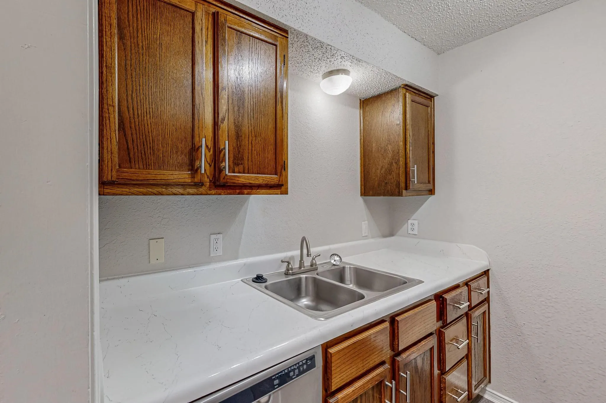 Wood-finish cabinetry with silver-tone pulls, dual basin stainless steel sink, white countertop, and stainless steel dishwasher
