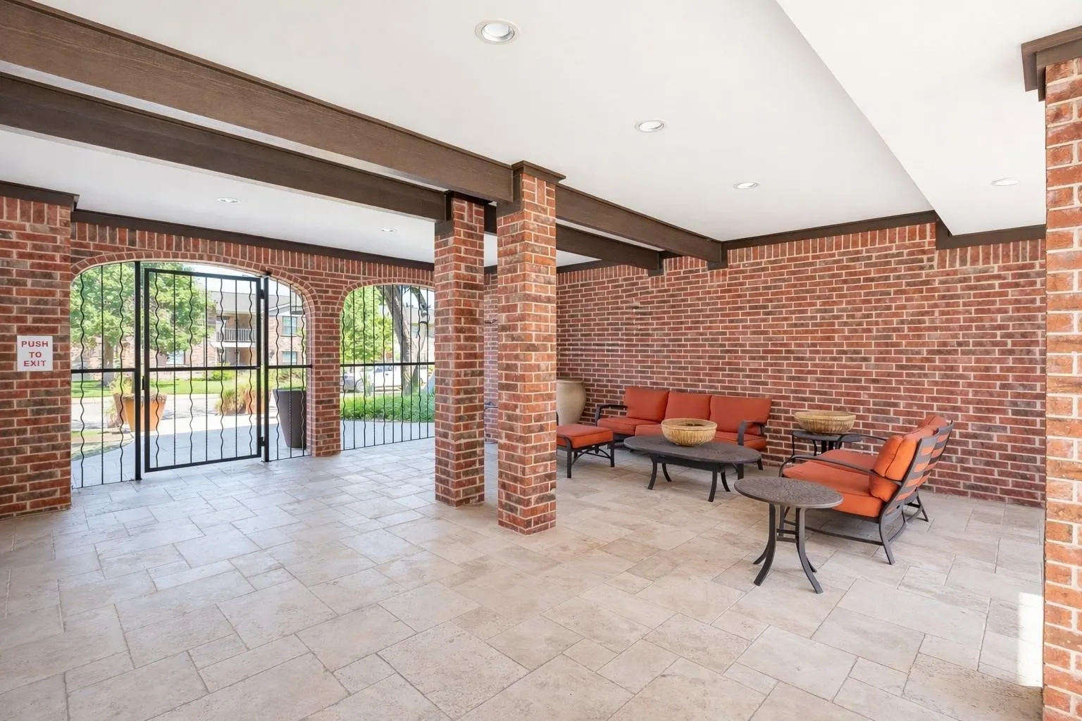 Covered outdoor gathering area featuring brick walls, arched openings, wood-finish beams, recessed lighting, and light-toned tile flooring
