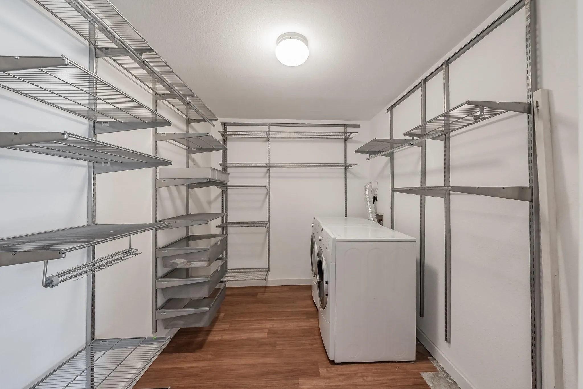 Dedicated laundry area featuring wood-finish flooring, white walls, overhead lighting, and extensive wire shelving with integrated drawers