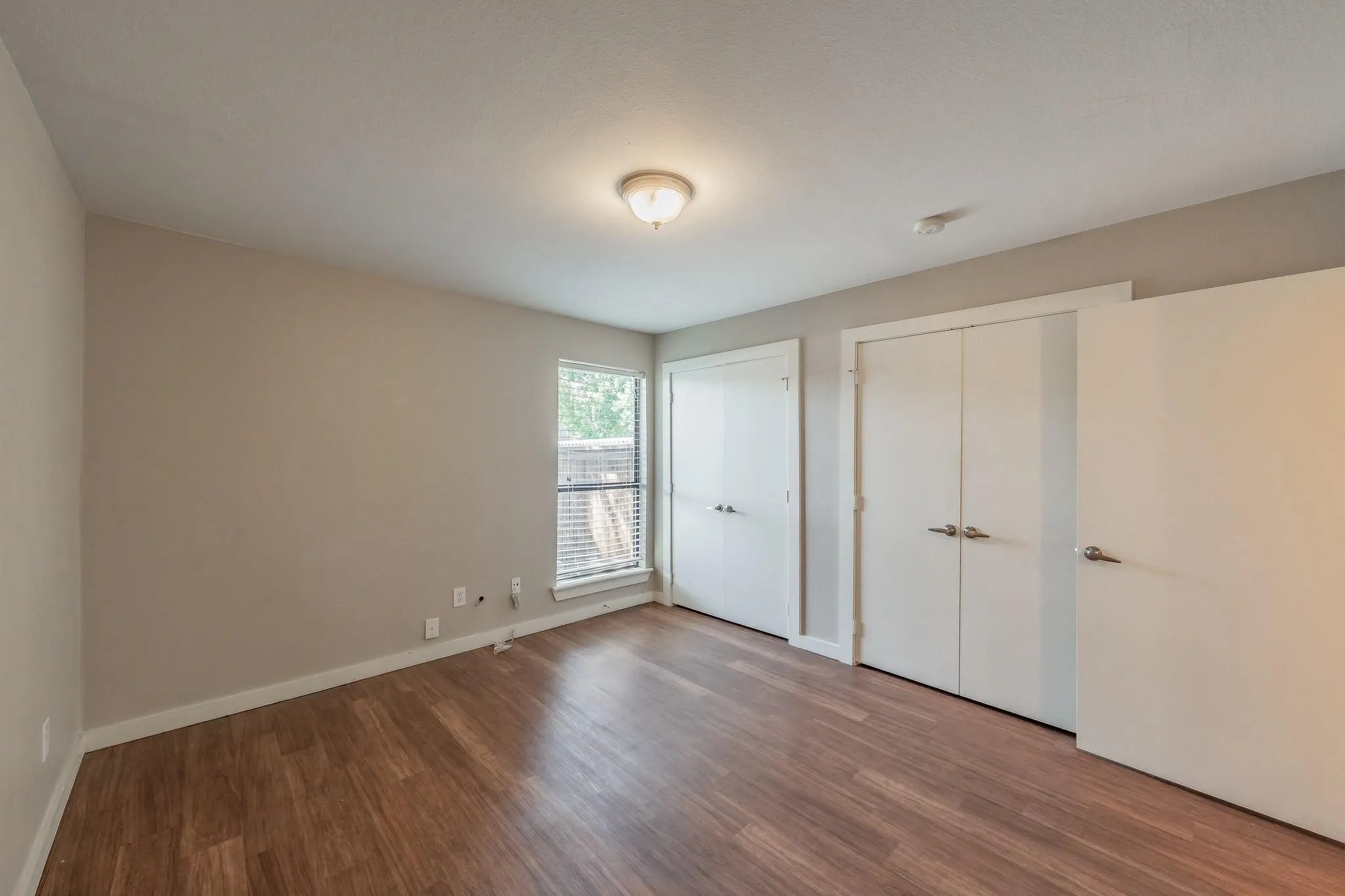 Wood-finish flooring, neutral wall paint, ceiling-mounted light fixture, window with blinds, and white doors
