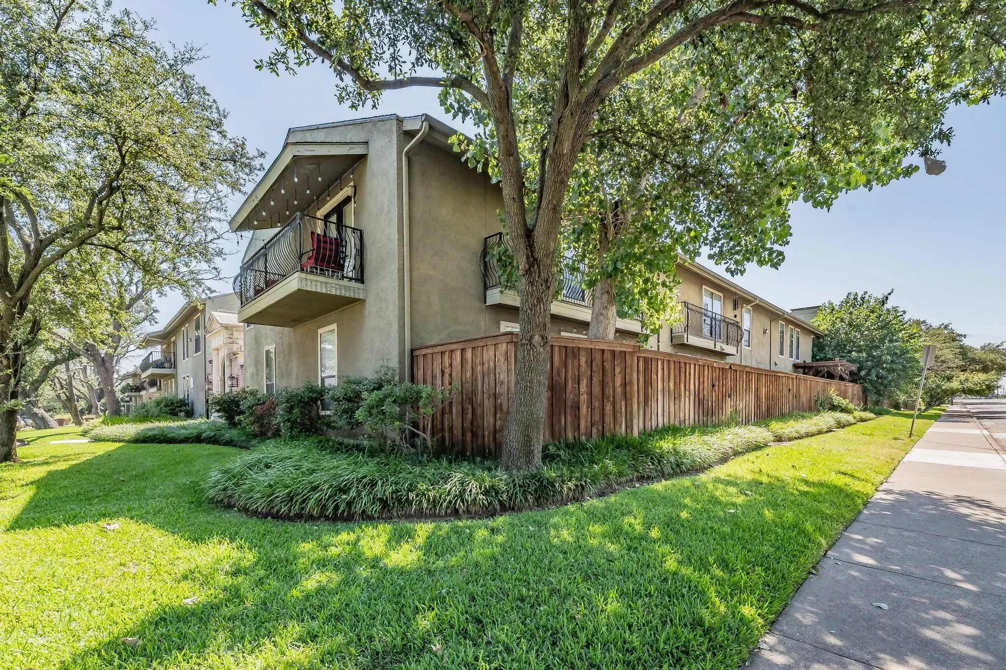 Stucco exterior featuring multiple balconies with black wrought iron railings, a wood privacy fence, and mature trees providing shade