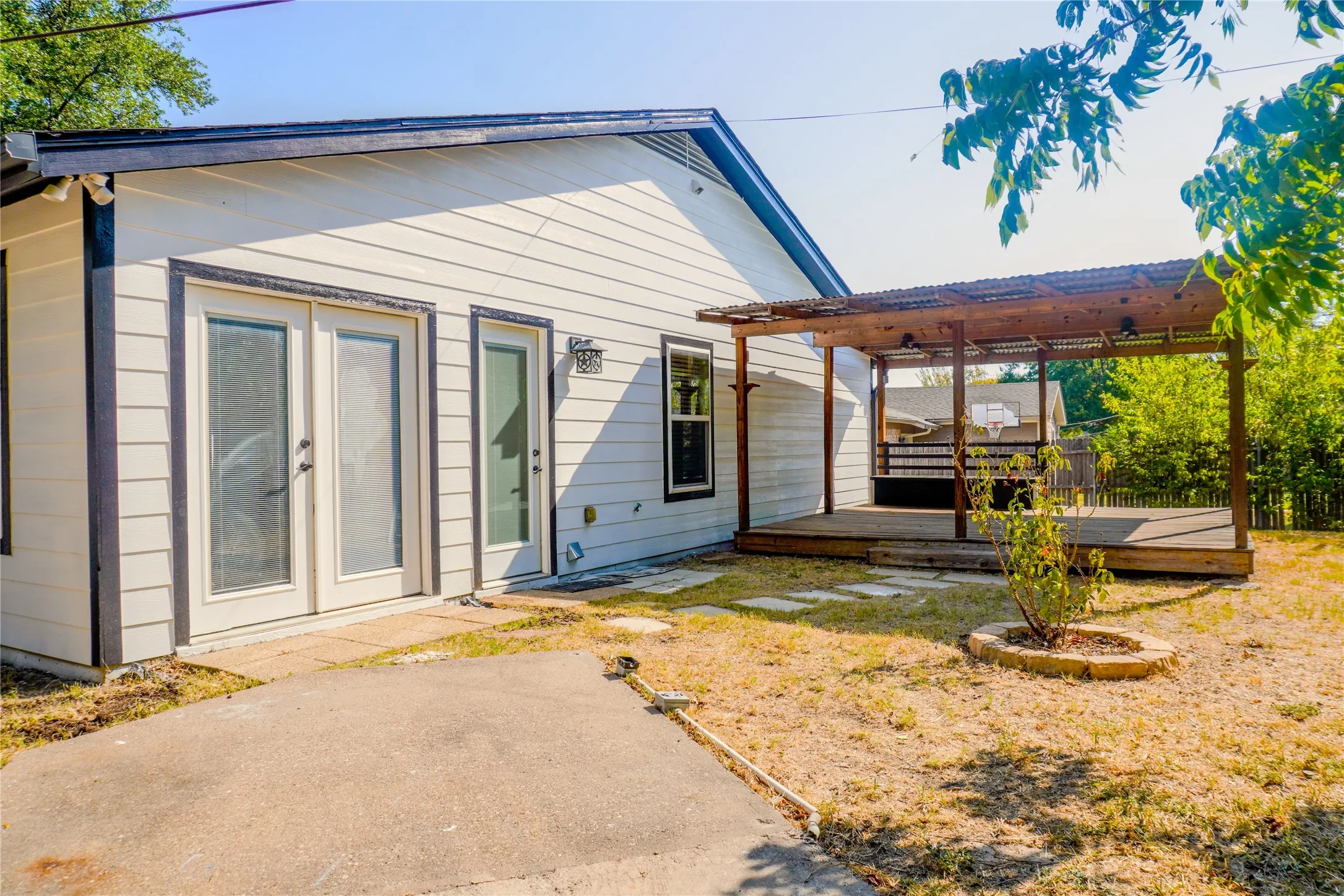 Exterior siding, double glass-paneled doors, wood deck with pergola, concrete patio area, and defined planting bed