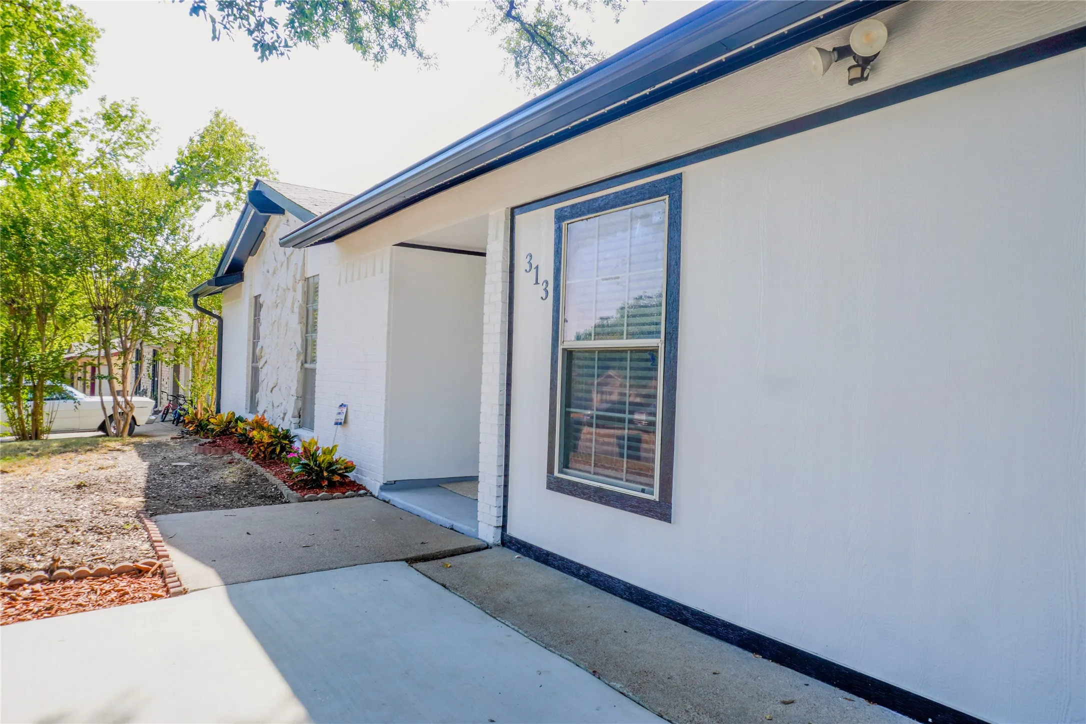 White exterior with dark trim accents, featuring a prominent window, a concrete pathway, and landscaped garden beds