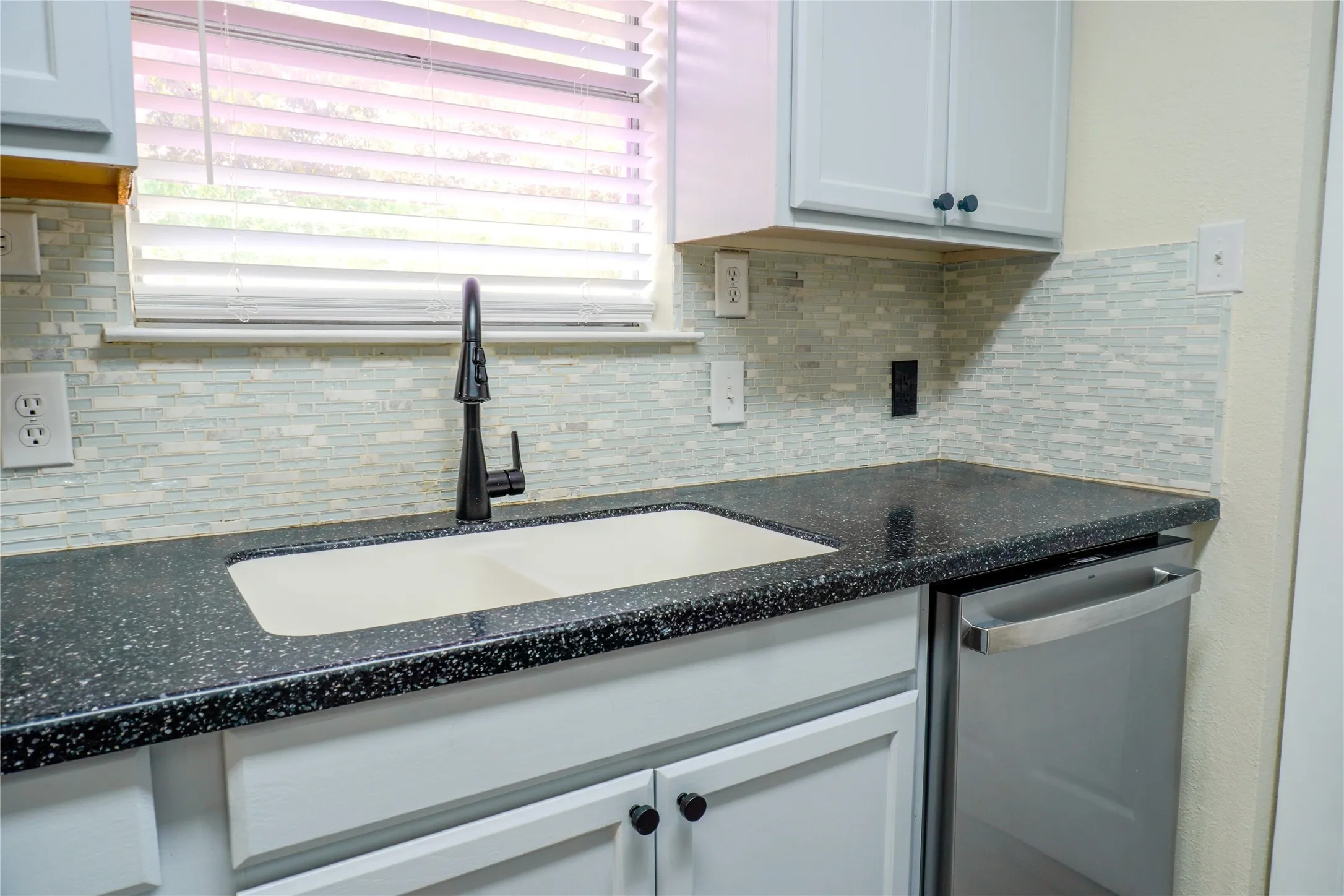 White shaker cabinetry with dark hardware, dark speckled countertops, undermount double-basin sink, matte black gooseneck faucet, and a light-toned mosaic tile backsplash