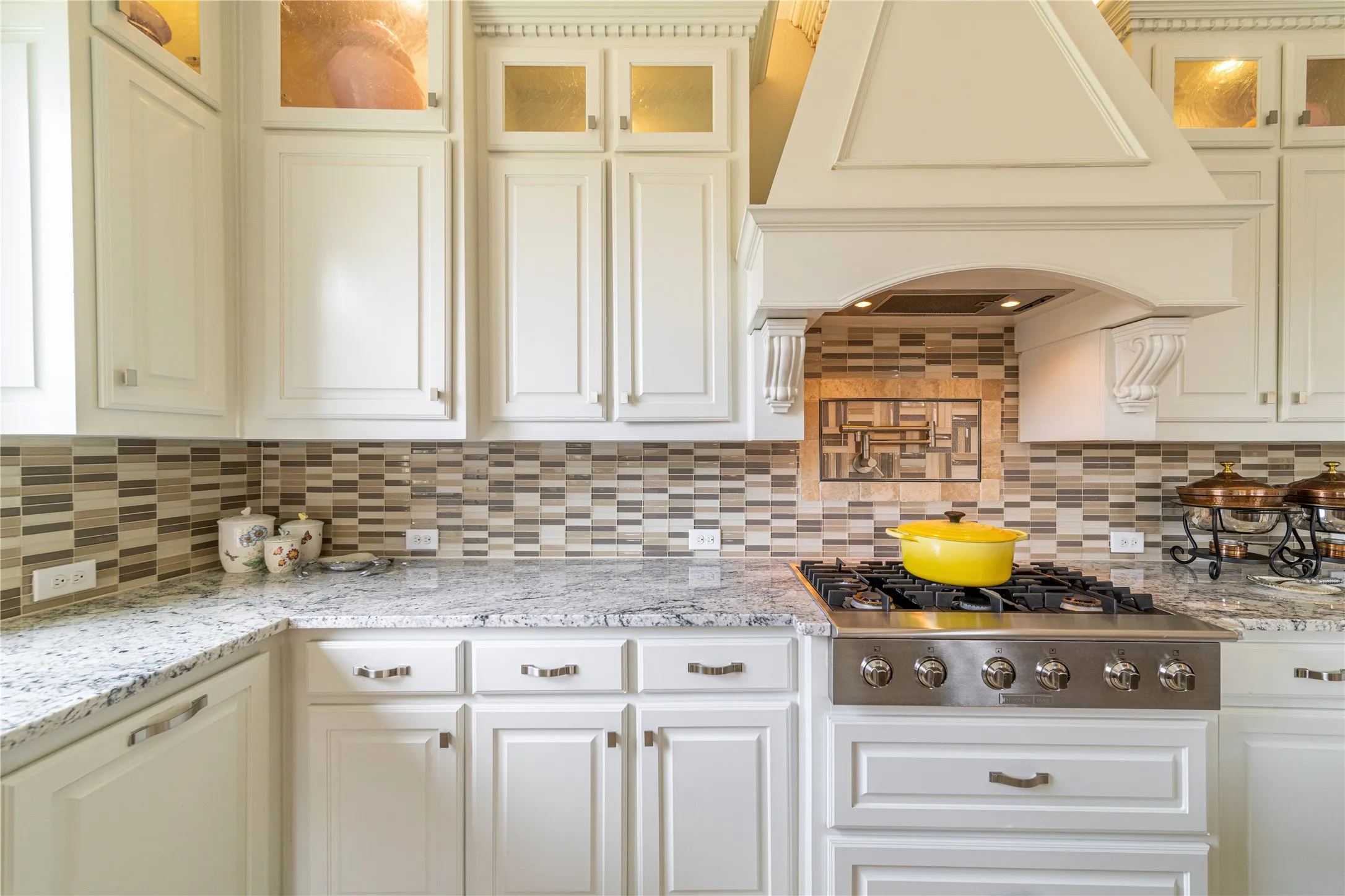 White custom cabinetry with crown molding, stainless steel gas range, custom range hood, mosaic tile backsplash, and granite countertops