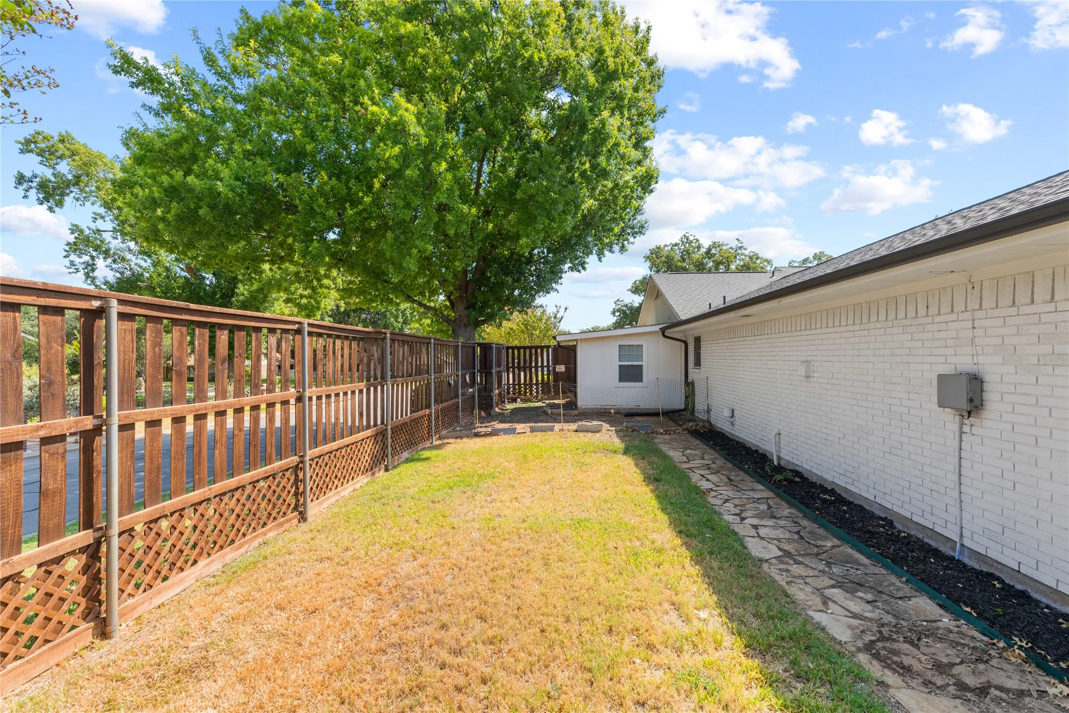Expansive backyard featuring a wood lattice fence, mature tree, white brick exterior, and a stone-paved walkway
