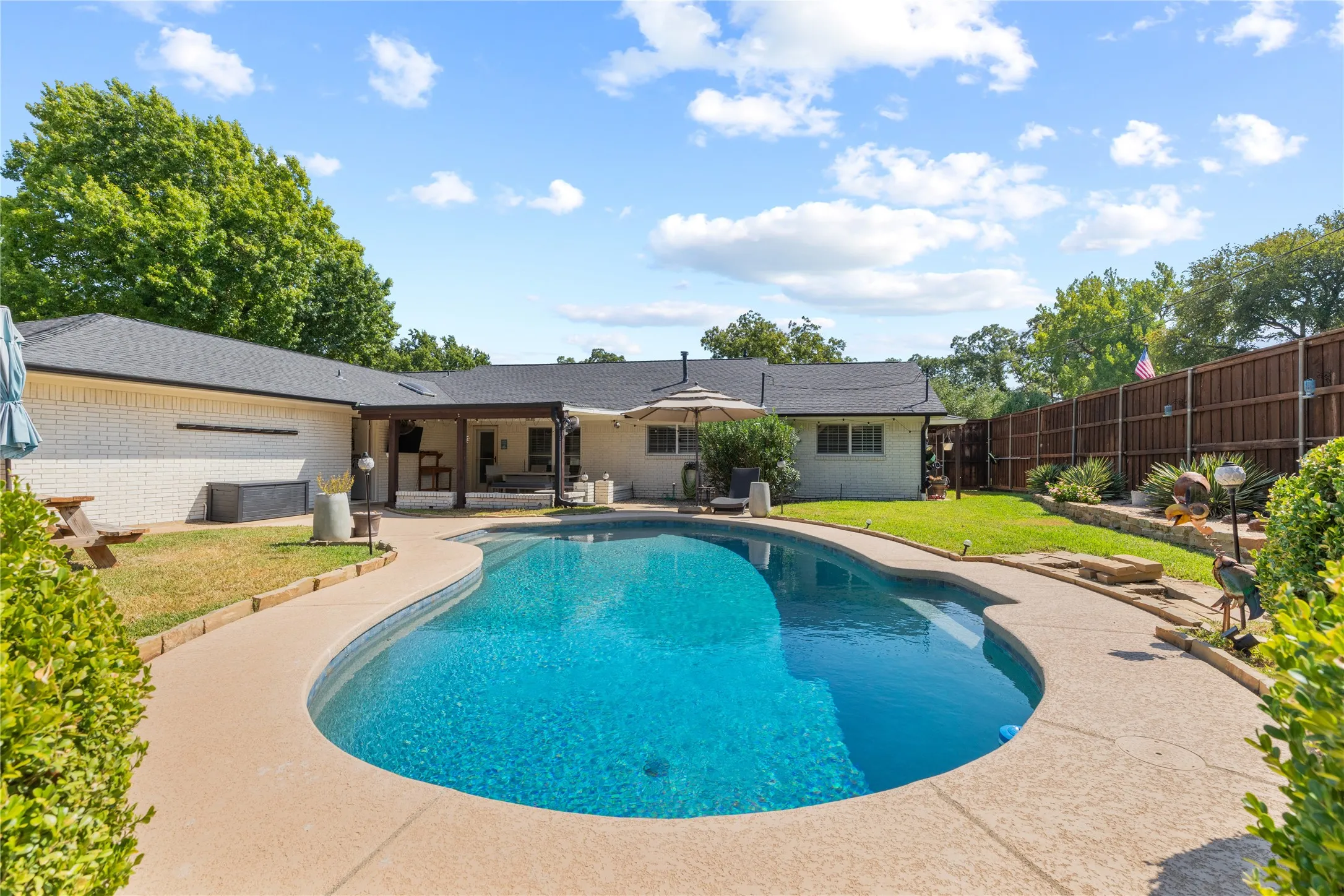 Curved inground swimming pool with a surrounding concrete deck, covered patio, and a privacy fence