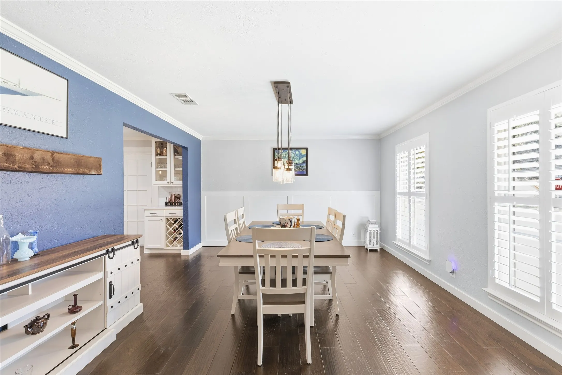 Hardwood flooring, crown molding, white wainscoting, white plantation shutters, and a kitchen pass-through with white cabinetry