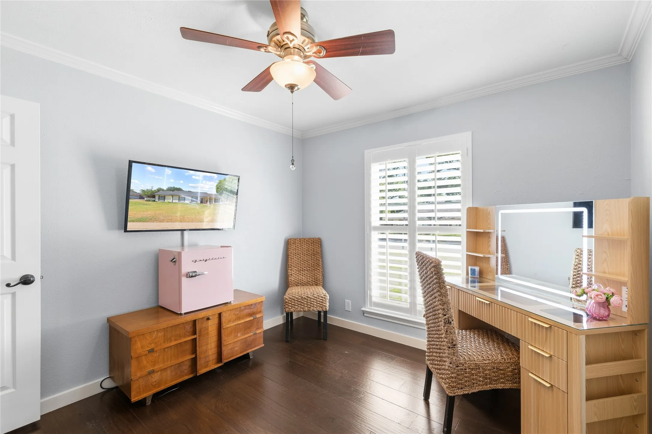 Dark wood-finish flooring, white crown molding, ceiling fan with integrated lighting, and window with white louvered shutters