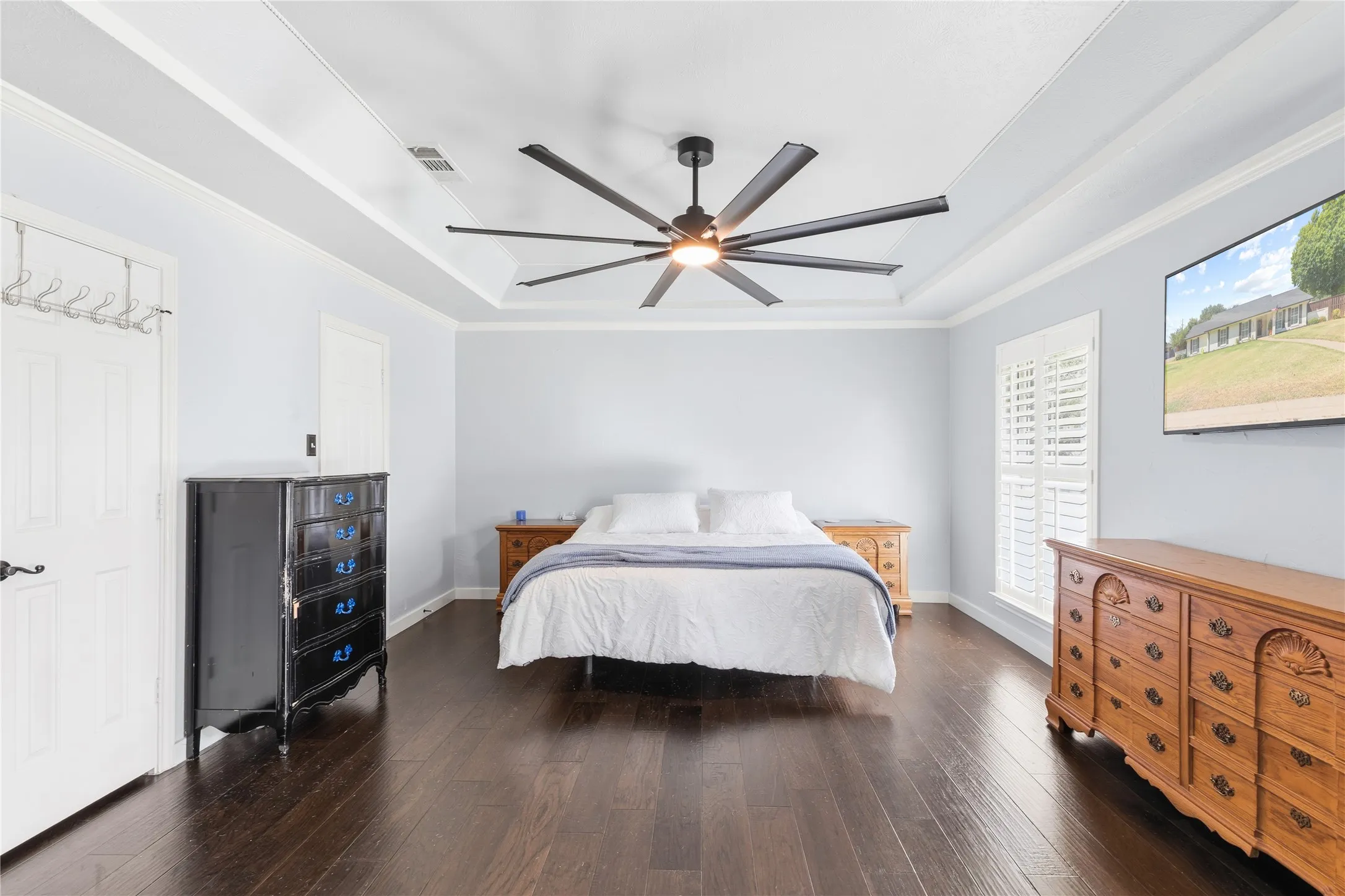 Tray ceiling with crown molding, dark wood-finish flooring, ceiling fan with integrated lighting, window with white plantation shutters, and a wall-mounted television