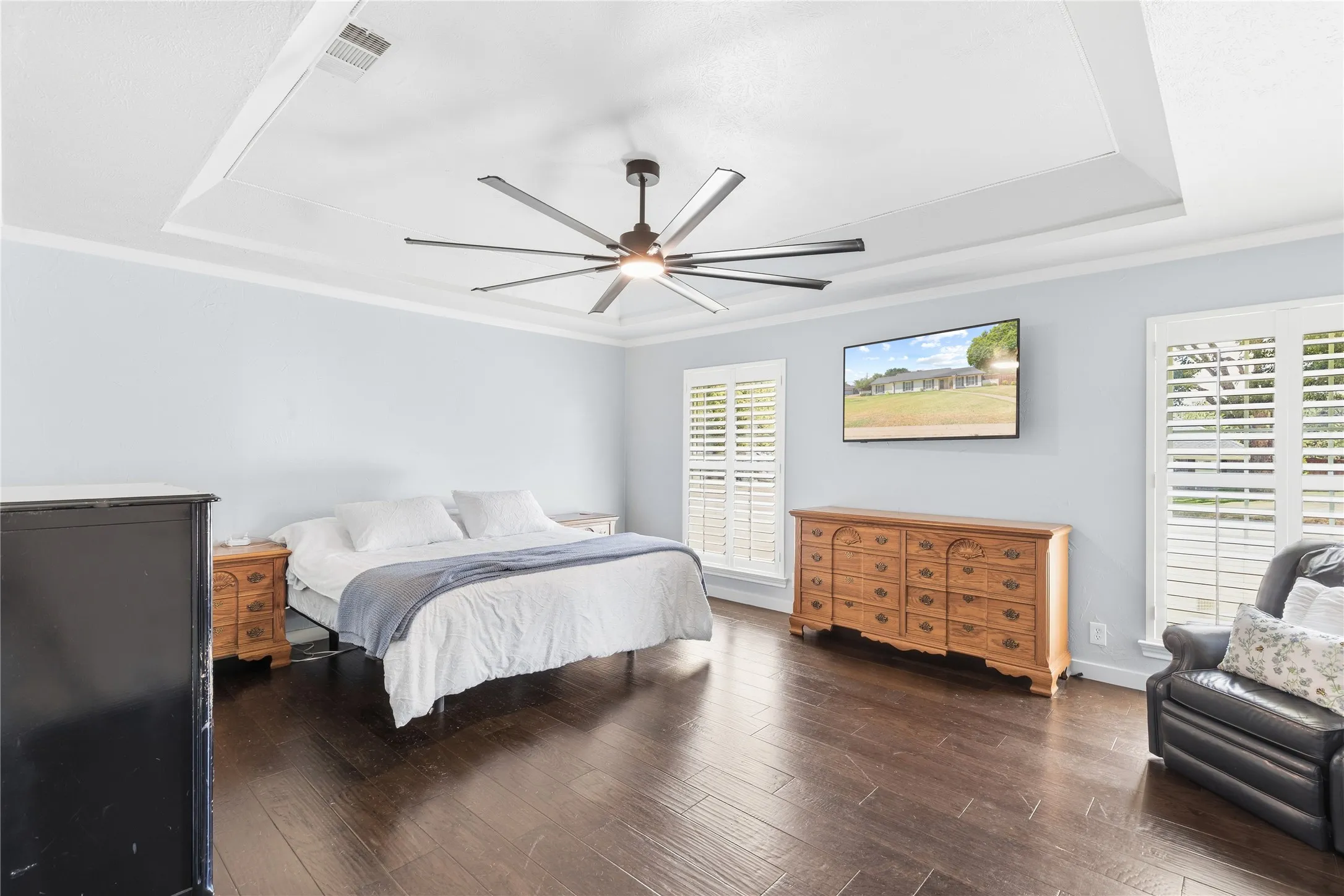 Tray ceiling with crown molding, wood-finish flooring, two windows with plantation shutters, and a ceiling fan