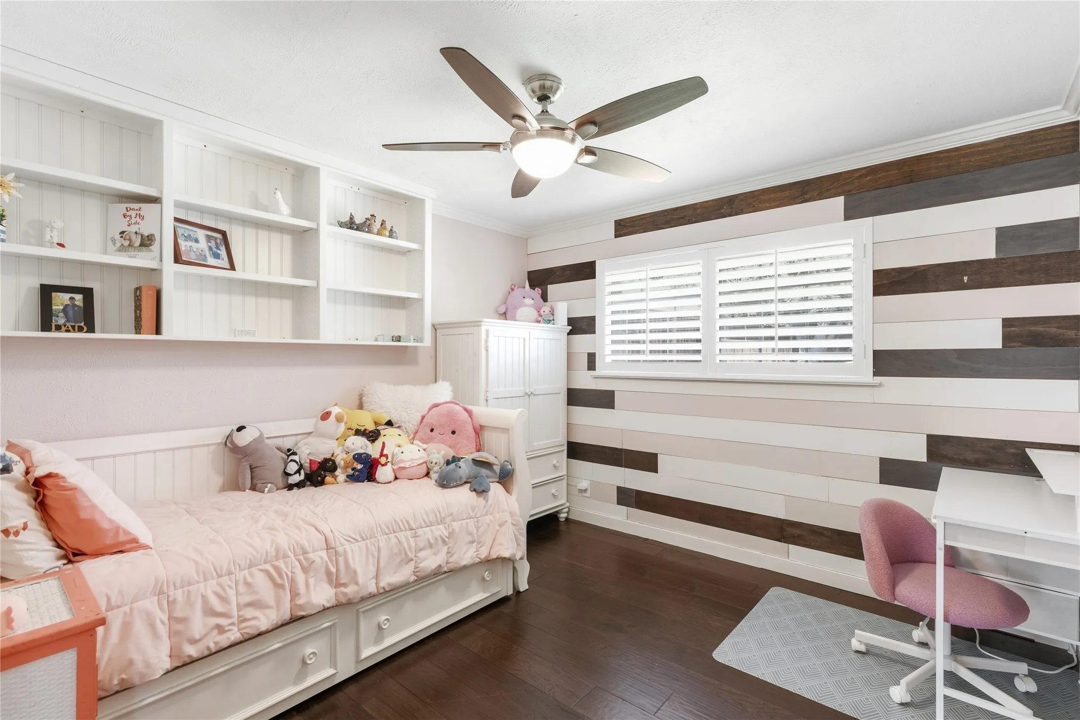 Built-in shelving, dark wood-finish flooring, ceiling fan with integrated lighting, window with white plantation shutters, and a decorative wood plank accent wall