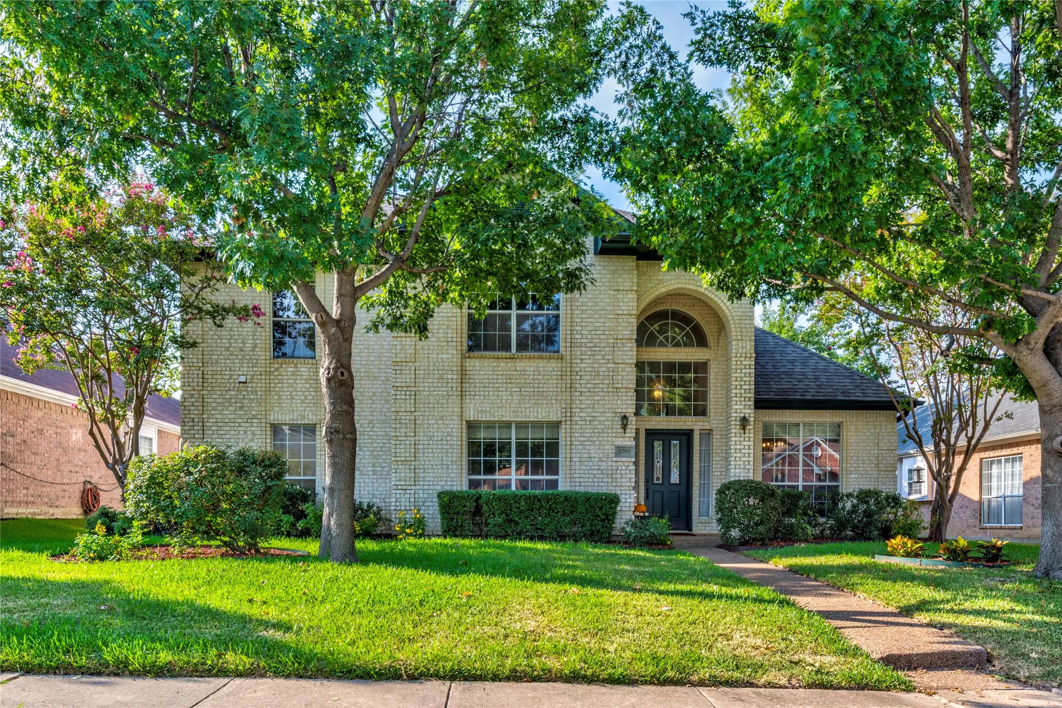 Brick exterior with beautiful mature trees, arched entry, dark green front door, and a concrete pathway....