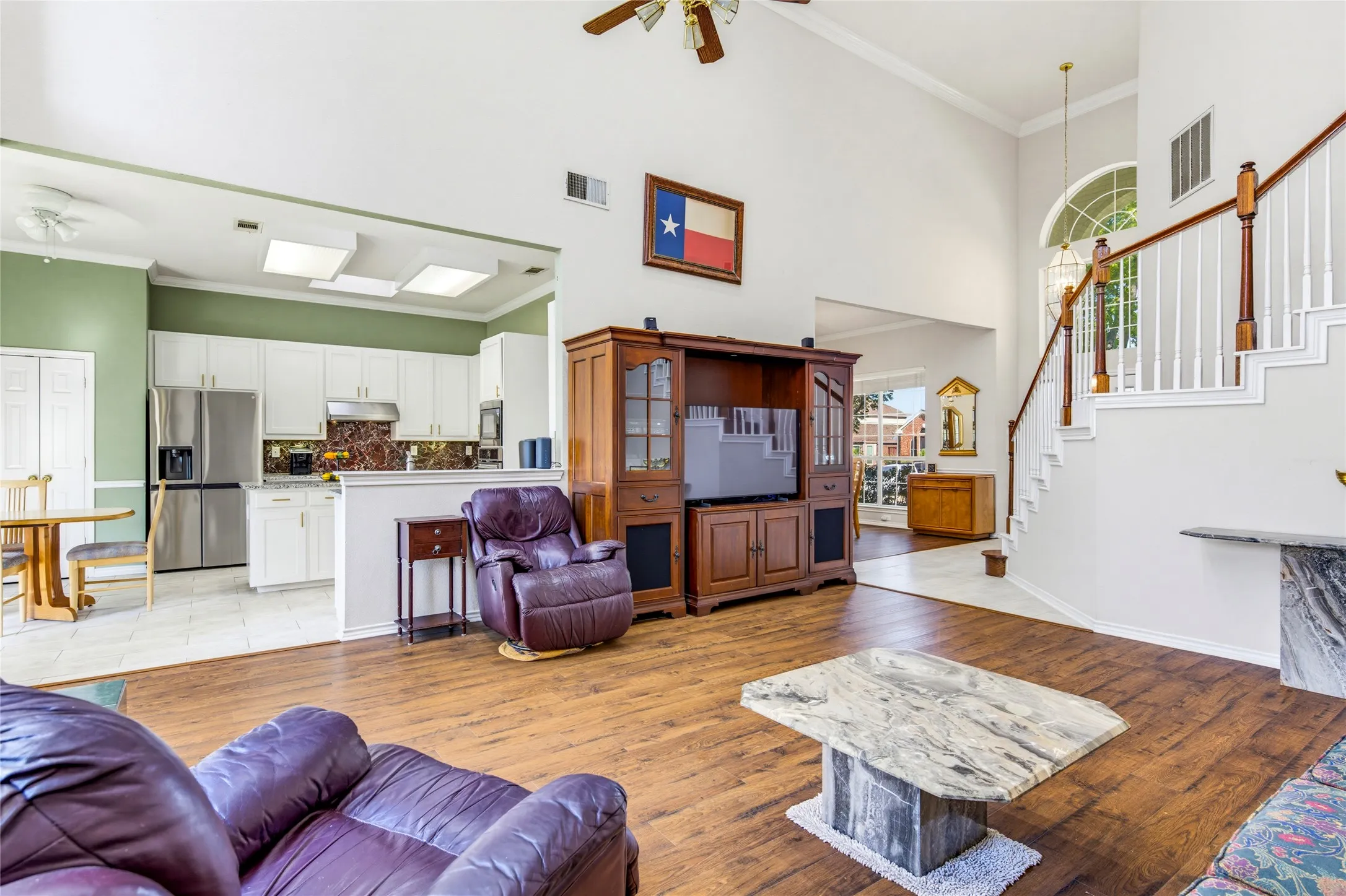 Vaulted ceiling with a ceiling fan, wood-finish laminate flooring, staircase with wood banister and white spindles, and kitchen with white cabinetry and stainless steel appliances, remodeled in 2023