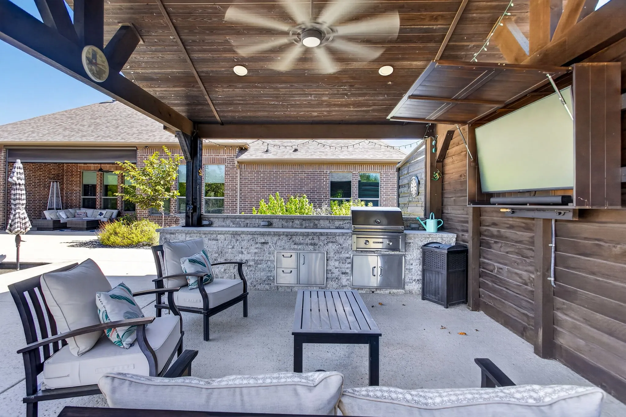 Covered patio featuring a built-in outdoor kitchen with grill, wood-paneled ceiling with recessed lighting and ceiling fan, and a wall-mounted entertainment cabinet