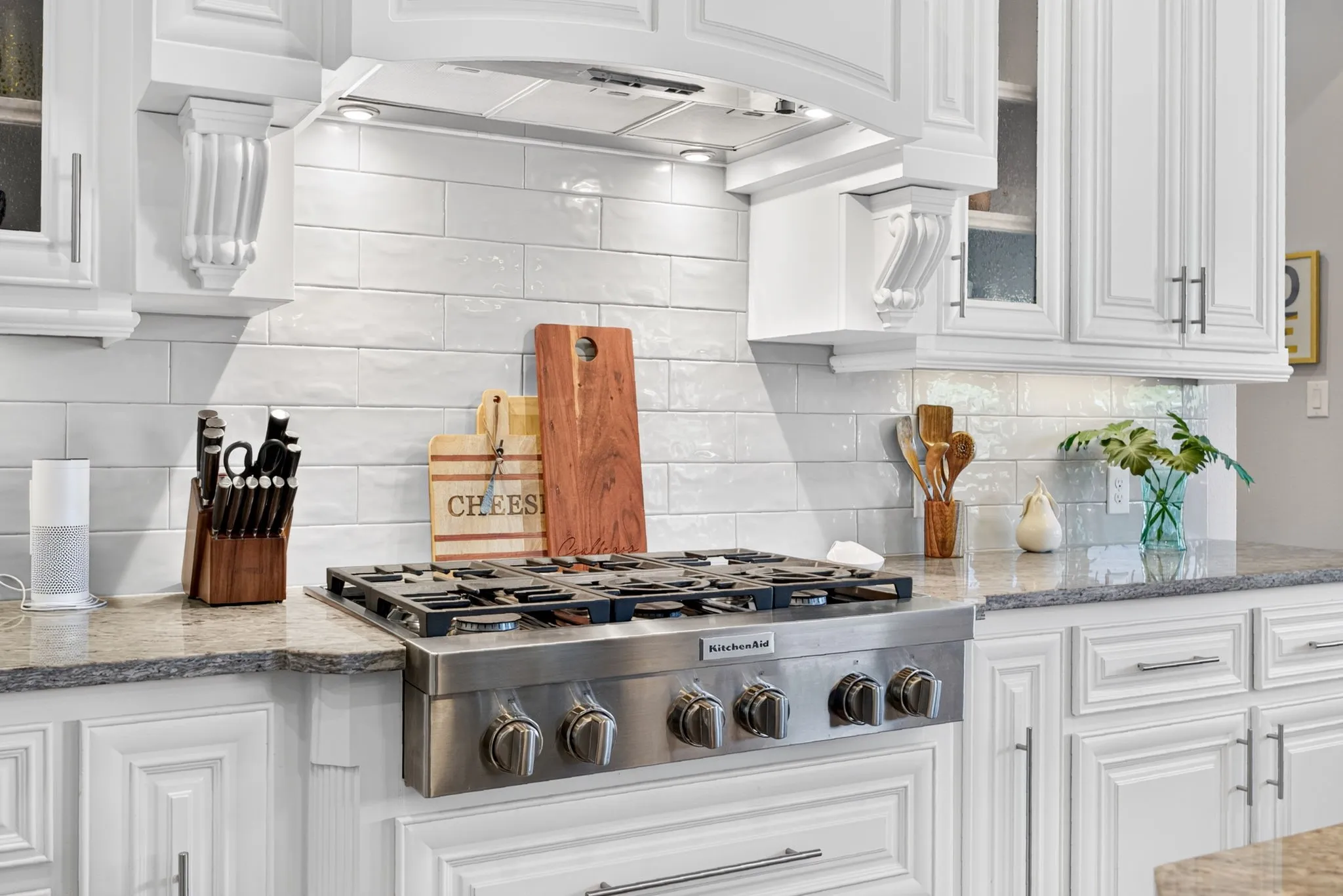 Built-in stainless steel gas cooktop, white subway tile backsplash, white cabinetry with decorative corbels, under-cabinet lighting, and stone countertops