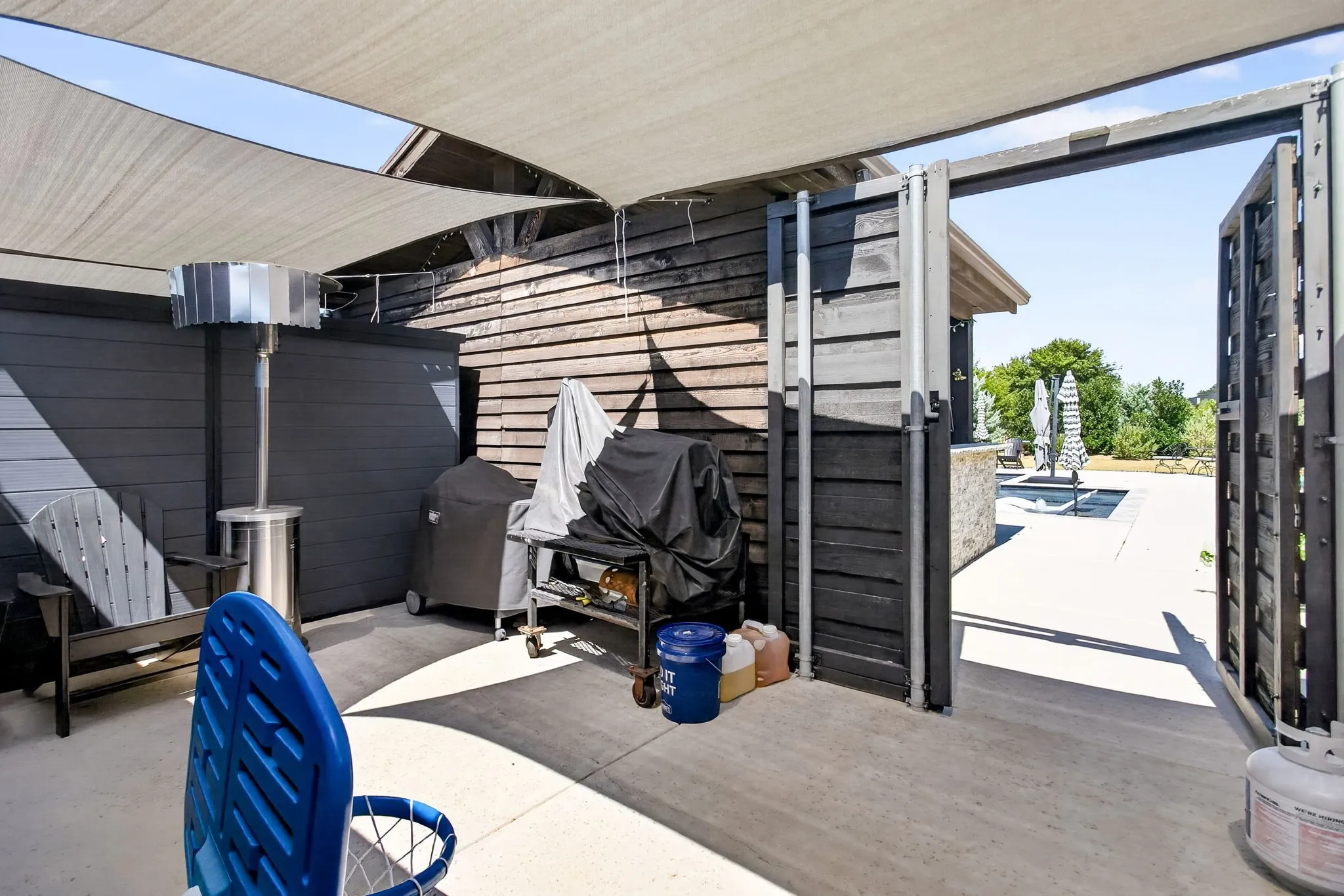 Paved patio area with a shade sail covering, featuring a privacy wall for patio and pool storage.