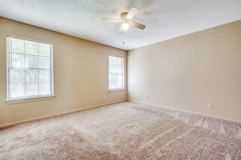 Spacious room featuring neutral carpeting, two large windows with blinds, a ceiling fan, and light-toned wall finishes