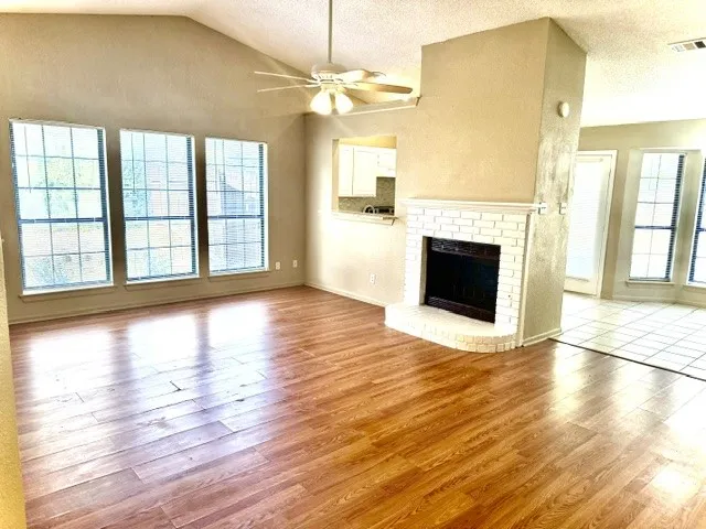 Vaulted ceiling with a ceiling fan, three large windows, wood-finish flooring, white brick fireplace, and a partial tile floor area