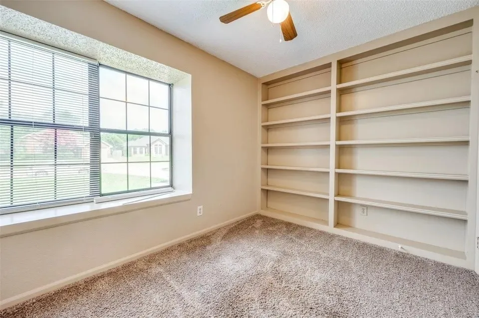 Built-in shelving, window with blinds, ceiling fan with light fixture, wall-to-wall carpeting