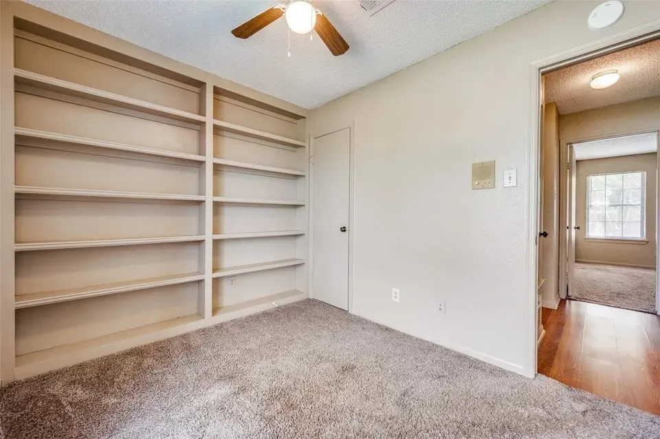 Built-in shelving, ceiling fan with light fixture, neutral carpeting, paneled interior doors, and wood-finish flooring in the adjacent space